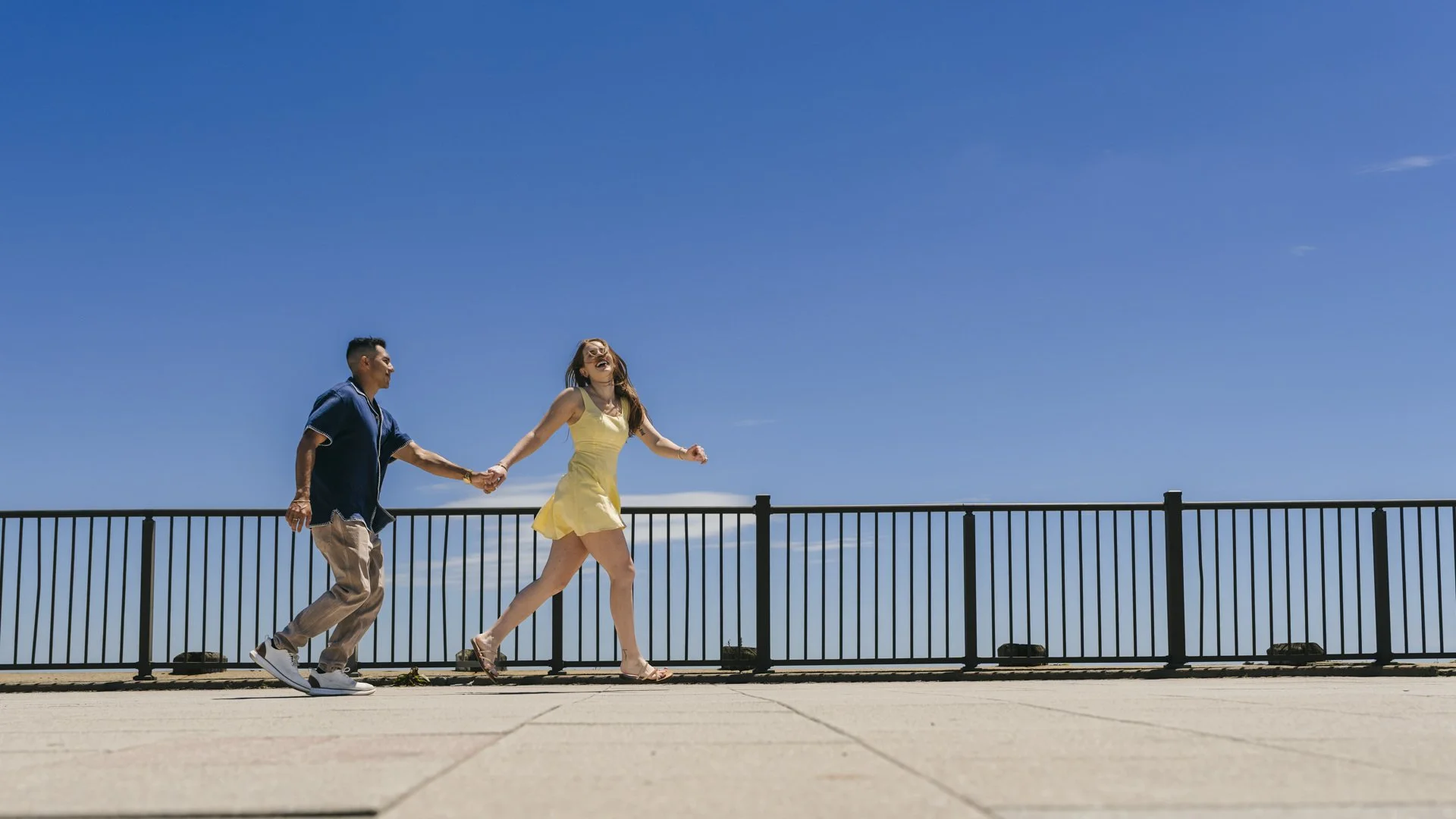 Couple laughing and holding hands along a waterfront railing beneath a blue sky