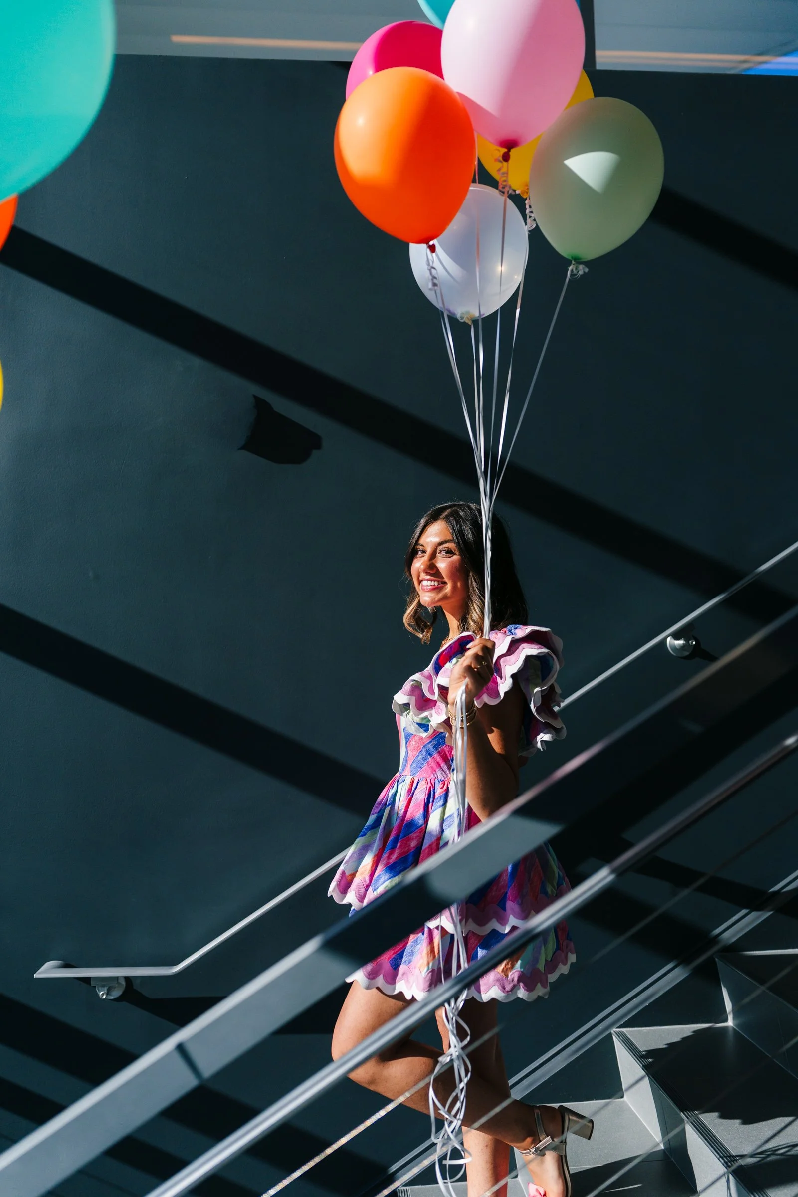 A sweet 16 girl in a colorful dress holding balloons on a staircase.