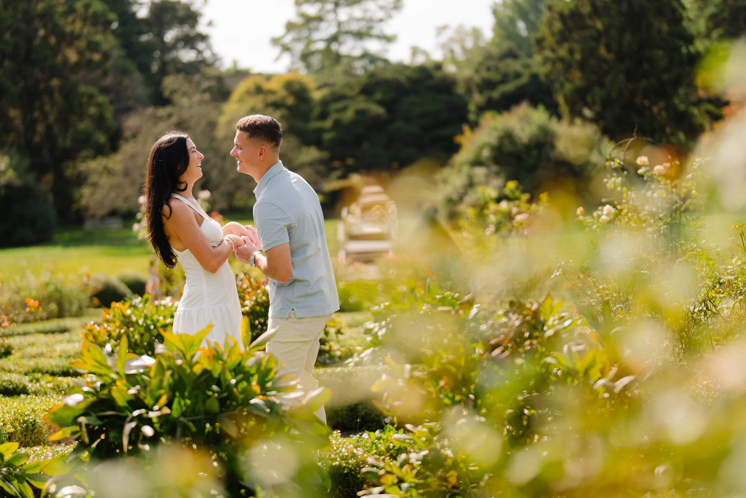 A couple smiling and holding hands in a garden with colorful flowers and lush greenery, enjoying a sunny day.
