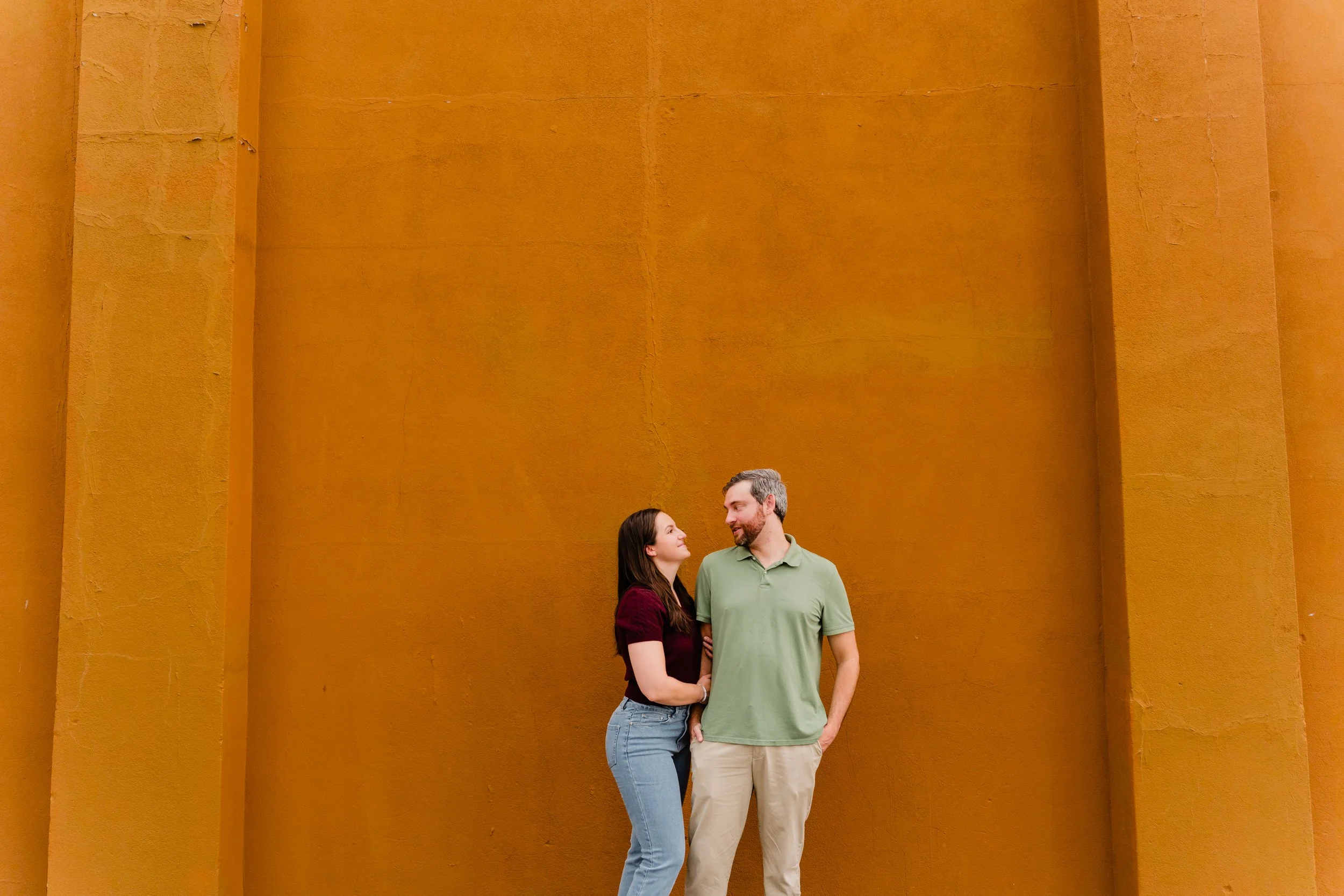 A couple stands close together, looking at each other and smiling, against a large, textured orange wall.