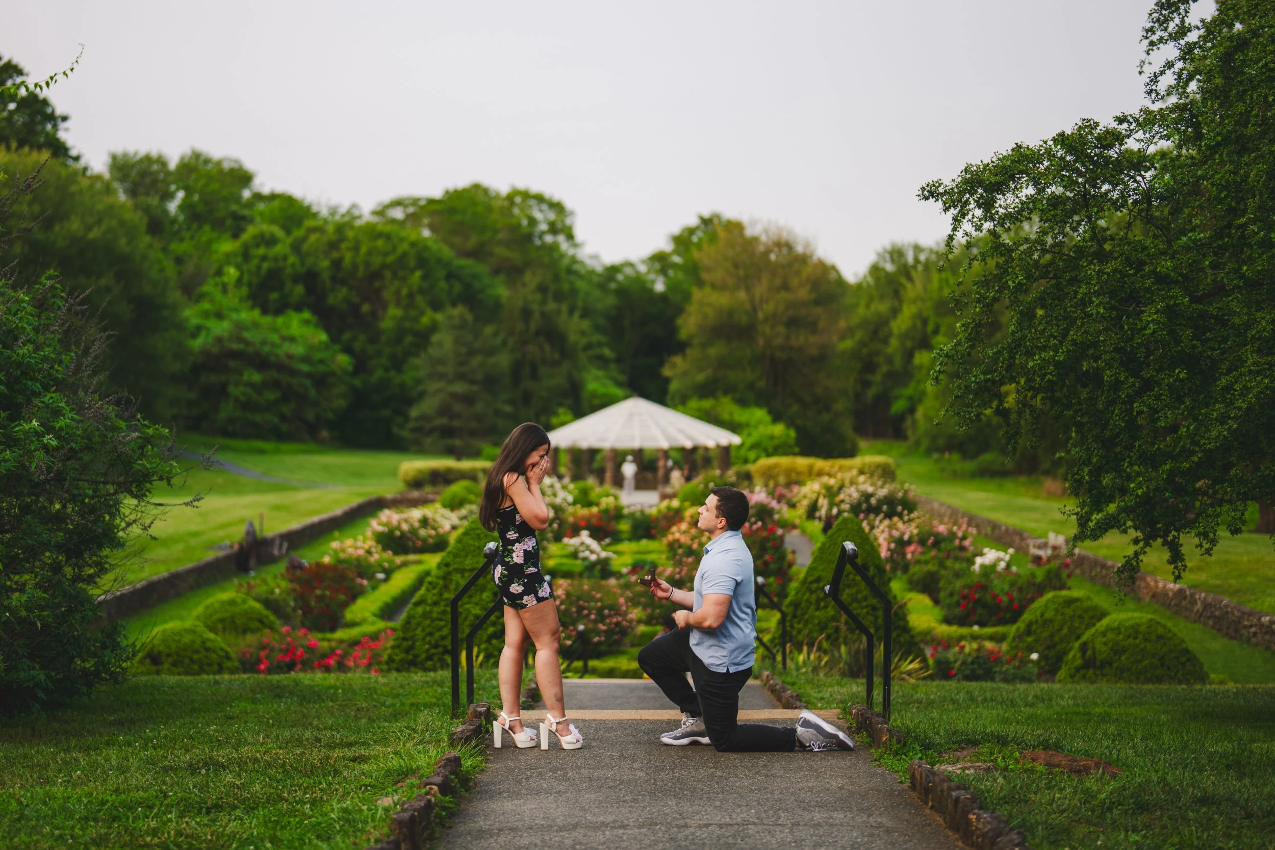 A man proposing to a woman on a garden path with flowers, greenery, and a gazebo in the background.