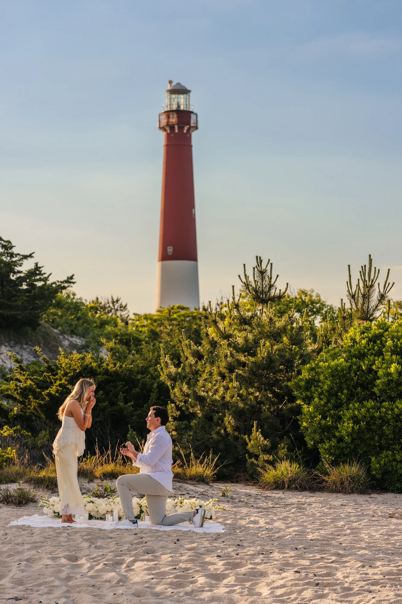 A surprise proposal on the sand with a red lighthouse in the background.