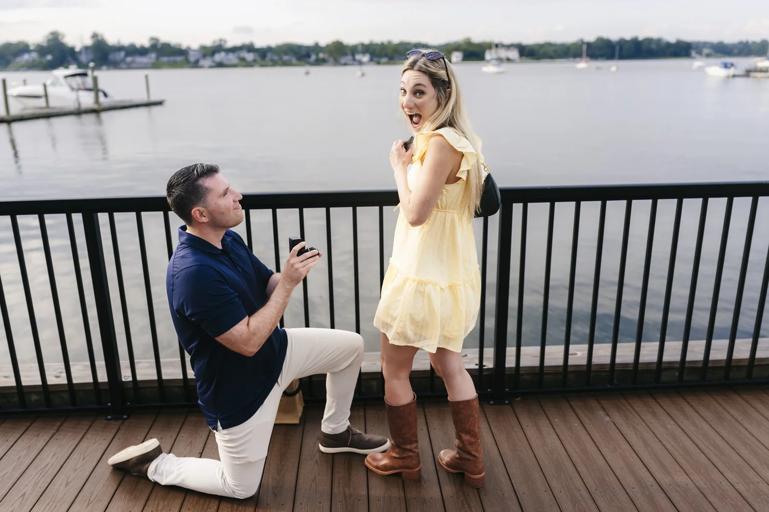 Man kneeling with a ring box as his partner reacts on a waterfront deck