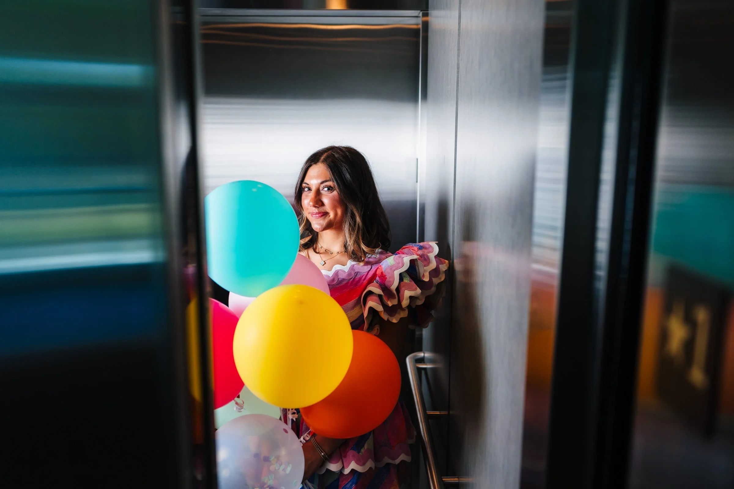 A sweet 16 girl in colorful clothing holding balloons stands in a narrow, metallic elevator shaft with reflections on the walls.