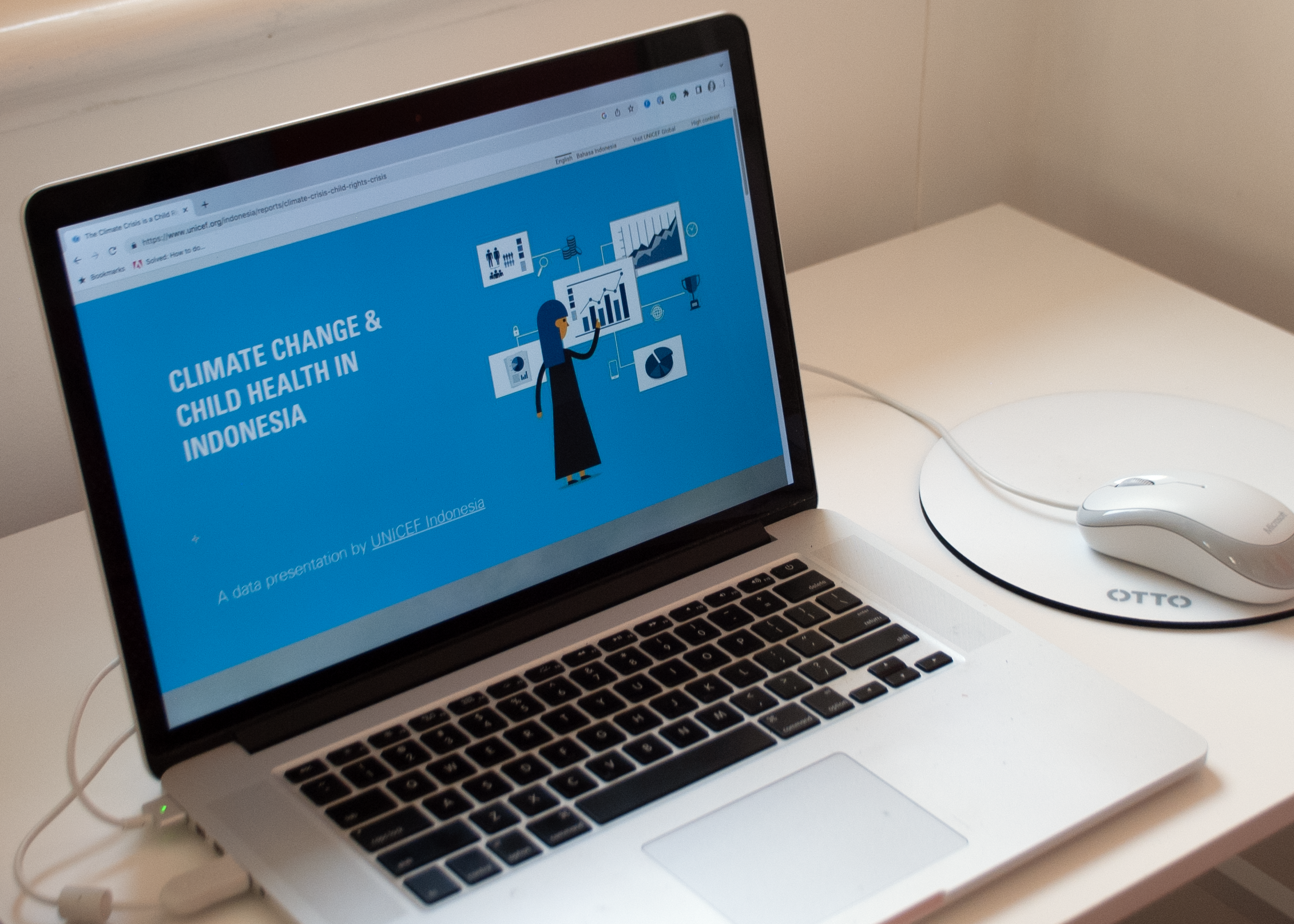 An opened laptop on a desk. The screen shows an illustration in light blue background with the texts Climate Change & Child Health in Indonesia on the left. The right side shows a woman facing several graphs.