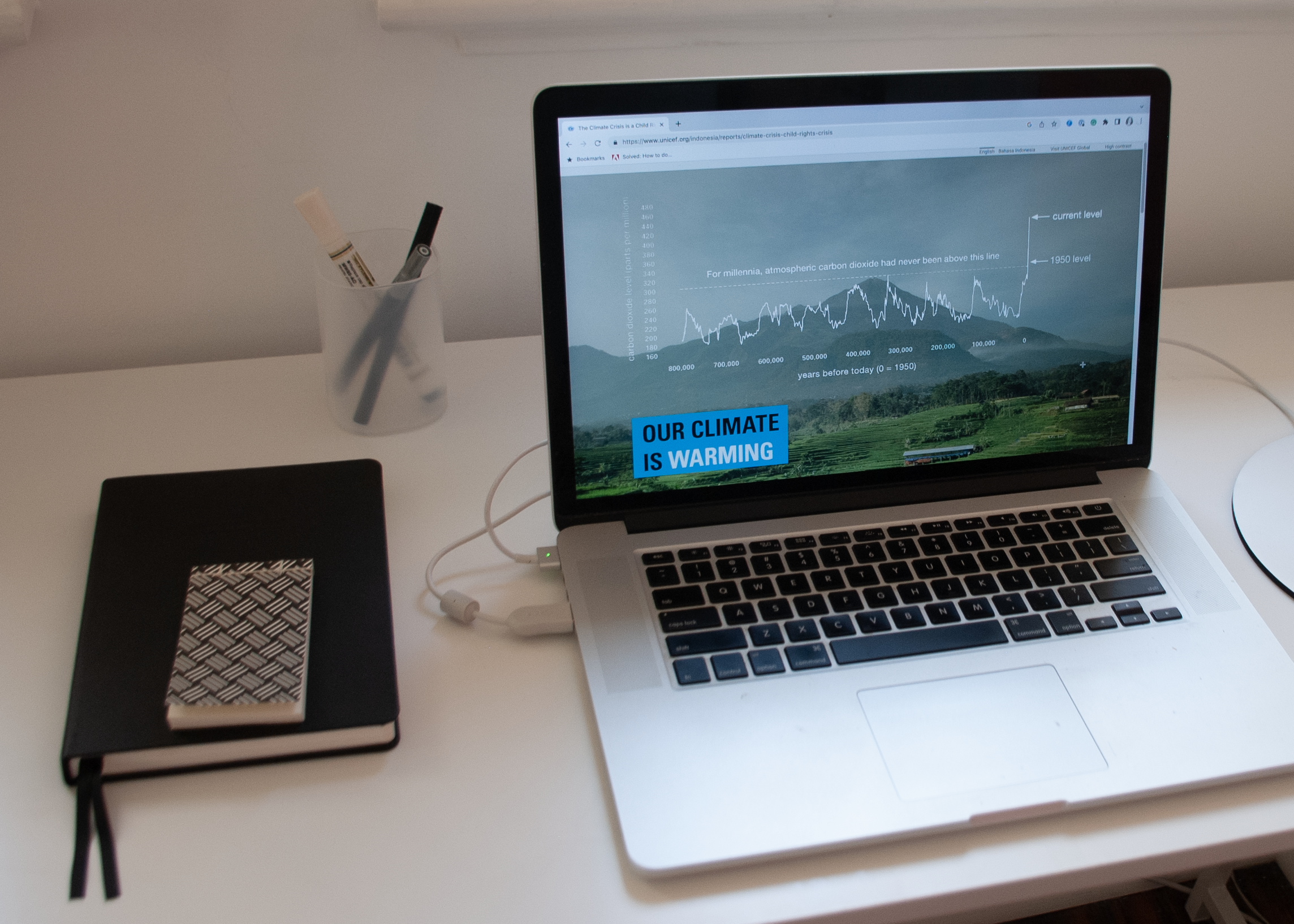 An opened laptop on a desk. The screen shows a graph with a background of a mountain and grass field. Lower left box shows the words Our Climate is Warming