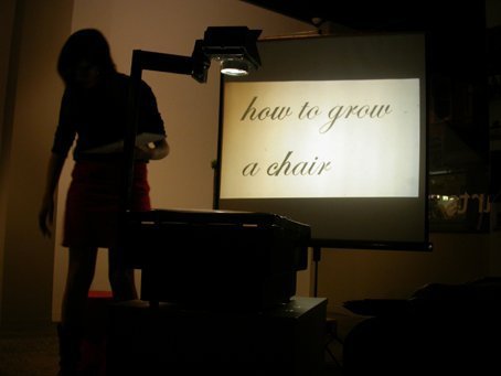A dimmed room. A woman’s silhouette is beside a white screen. The screen shows a cursive text of ‘how to grow a chair’