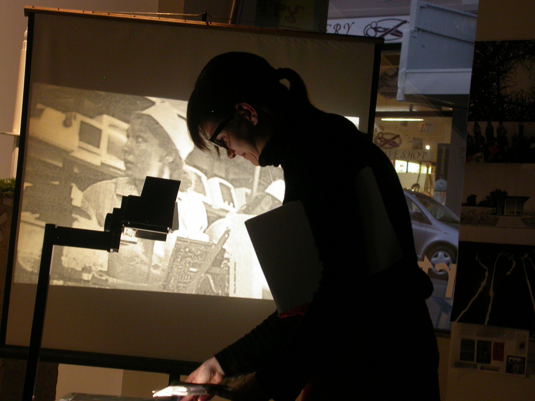 A dimmed room. A woman working the projector out. The white screen shows a woman looking at something on her right side.