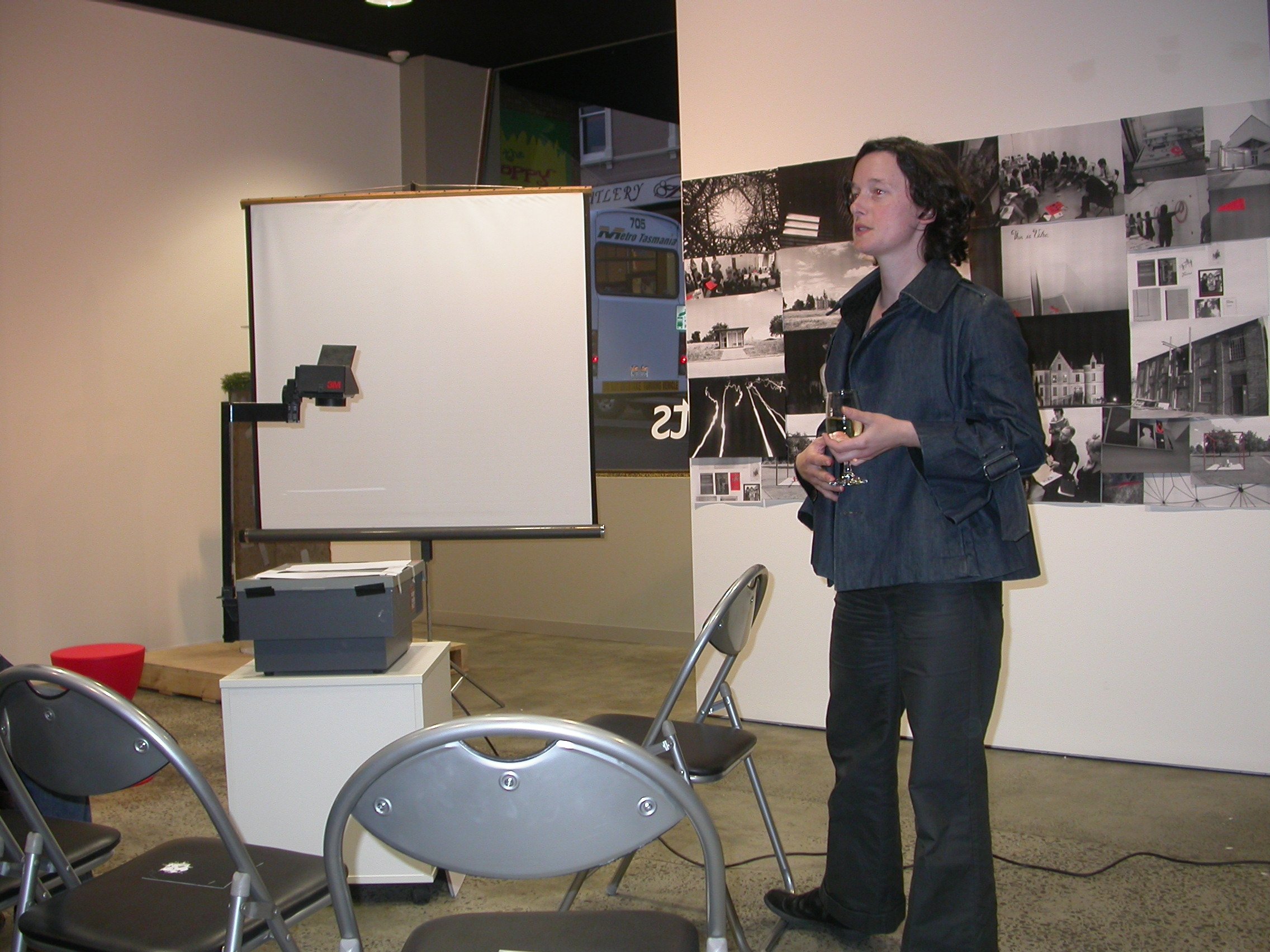 A woman speaking in front with a wine glass in her hand. She is beside the projector. She is facing the audience.