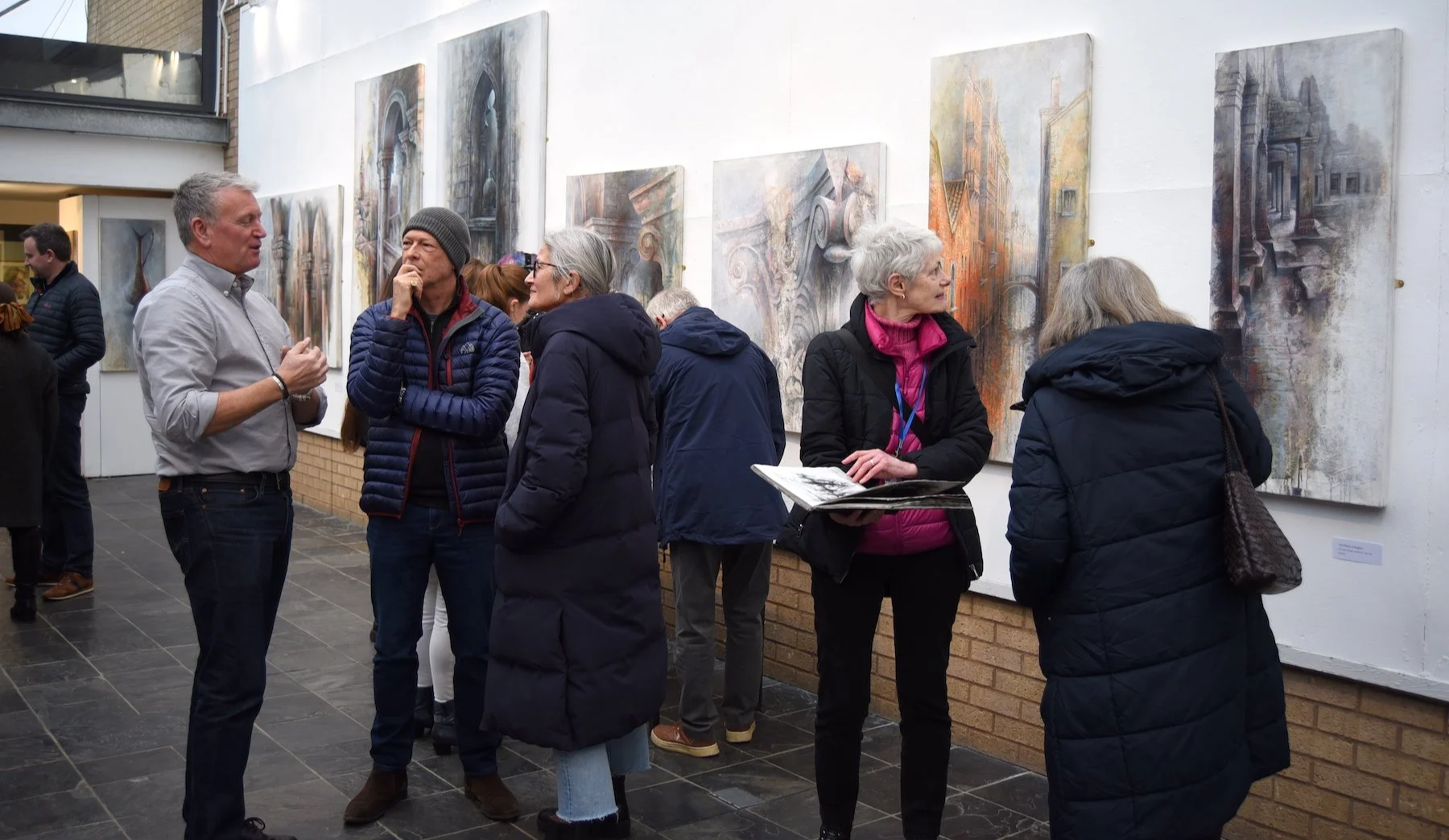Artist Ian Murphy in the gallery with his paintings on the wall behind. Ian is talking to some visitors, while others look at sketchbooks and the paintings on display.