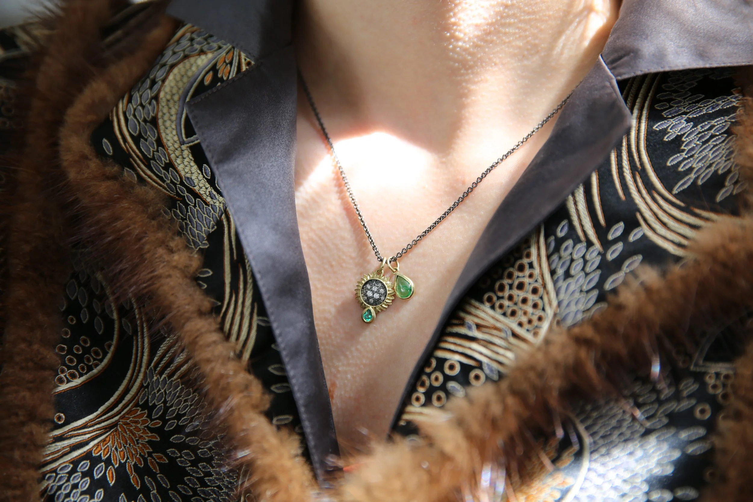 Medium sunflower pendant with emerald & tsavorite charm on oxi silver chain, white female model. Handmade fine art jewelry by Rebecca Myers Design.