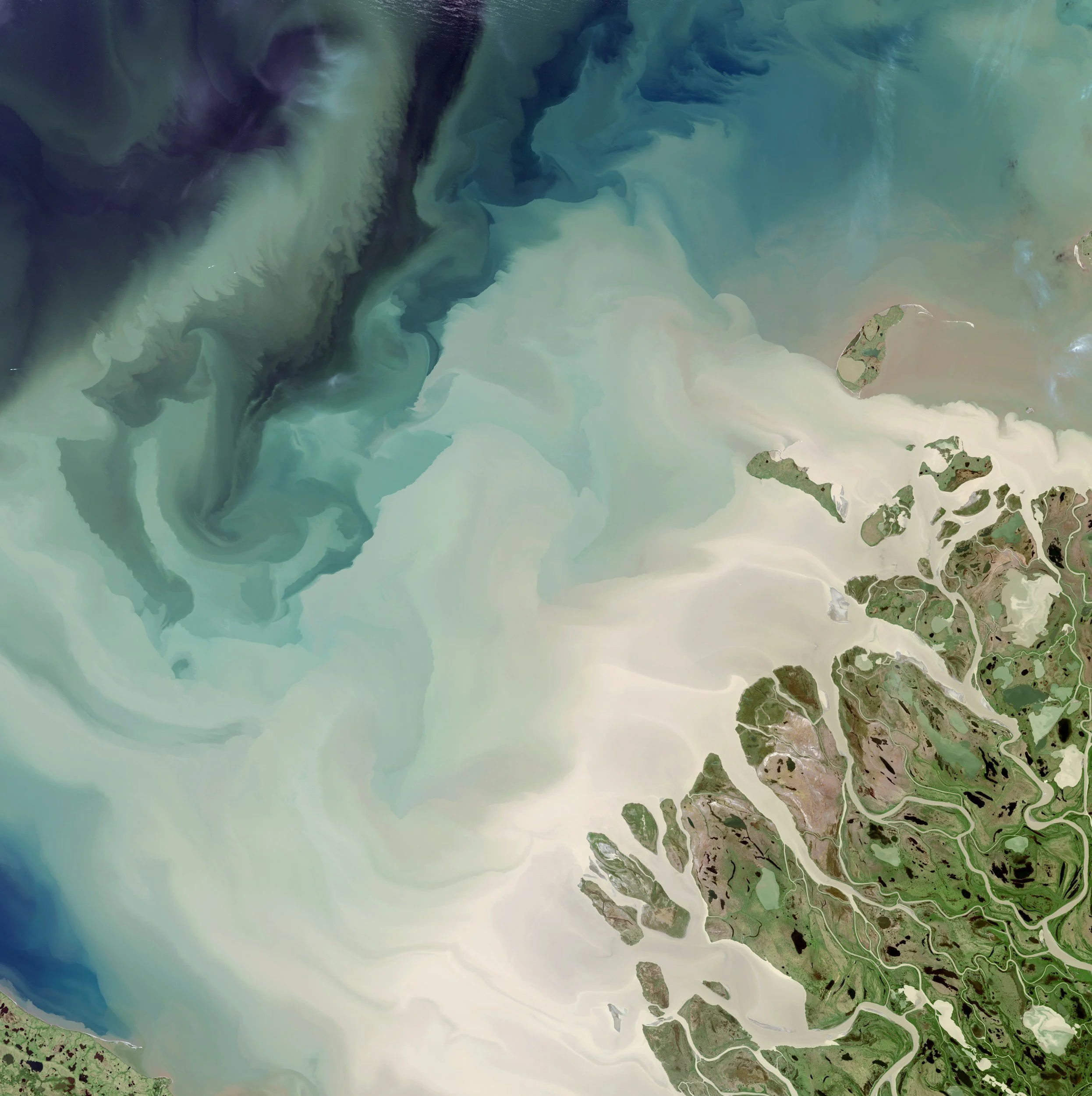 Aerial view of a coastal area with a large bay or inlet, showing green land, sandy beaches, and water with varying shades of blue and green, indicating different depths and possibly shallow areas near the shoreline.