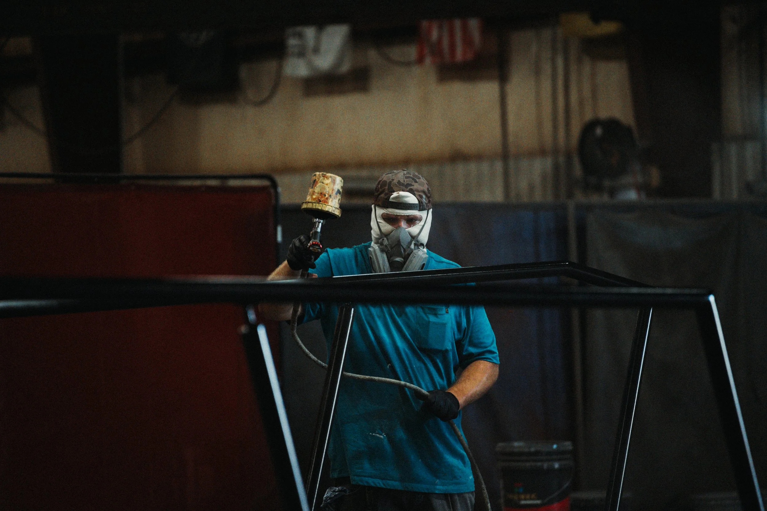A worker wearing a face mask, gloves, and a camouflage cap is using a spray gun to paint a surface in an industrial setting.