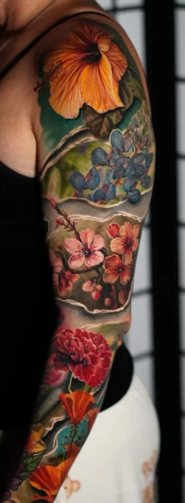 Close-up of a person's arm with colorful floral and nature-themed tattoos, including flowers, leaves, and butterflies, against a background of a black screen and a white panel.