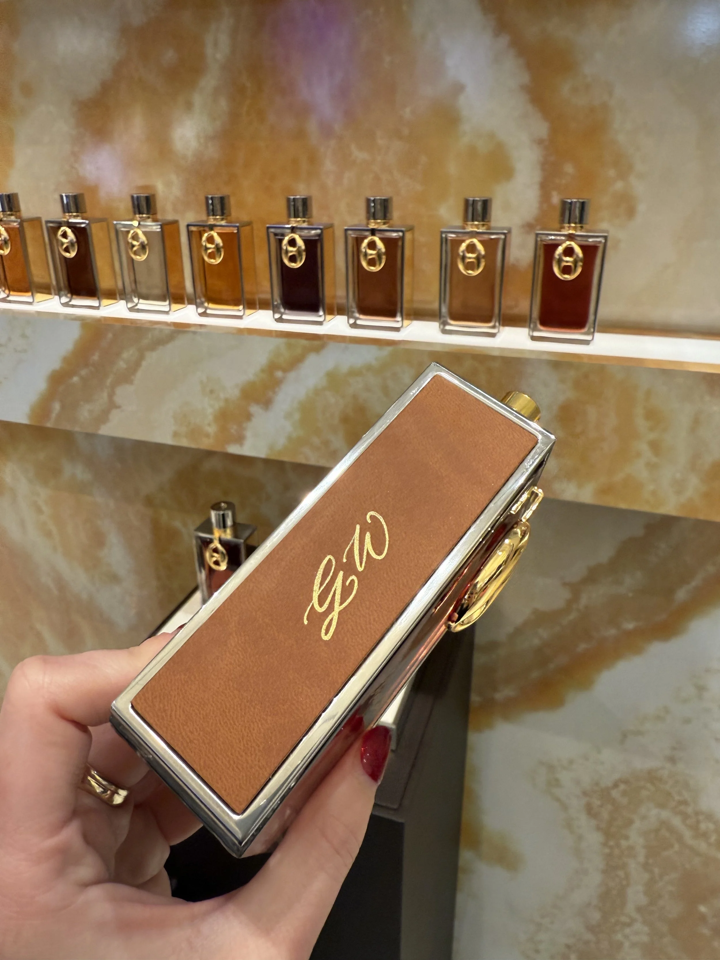 A hand holding a brown Gucci clutch bag in front of a display of perfume bottles in a store.