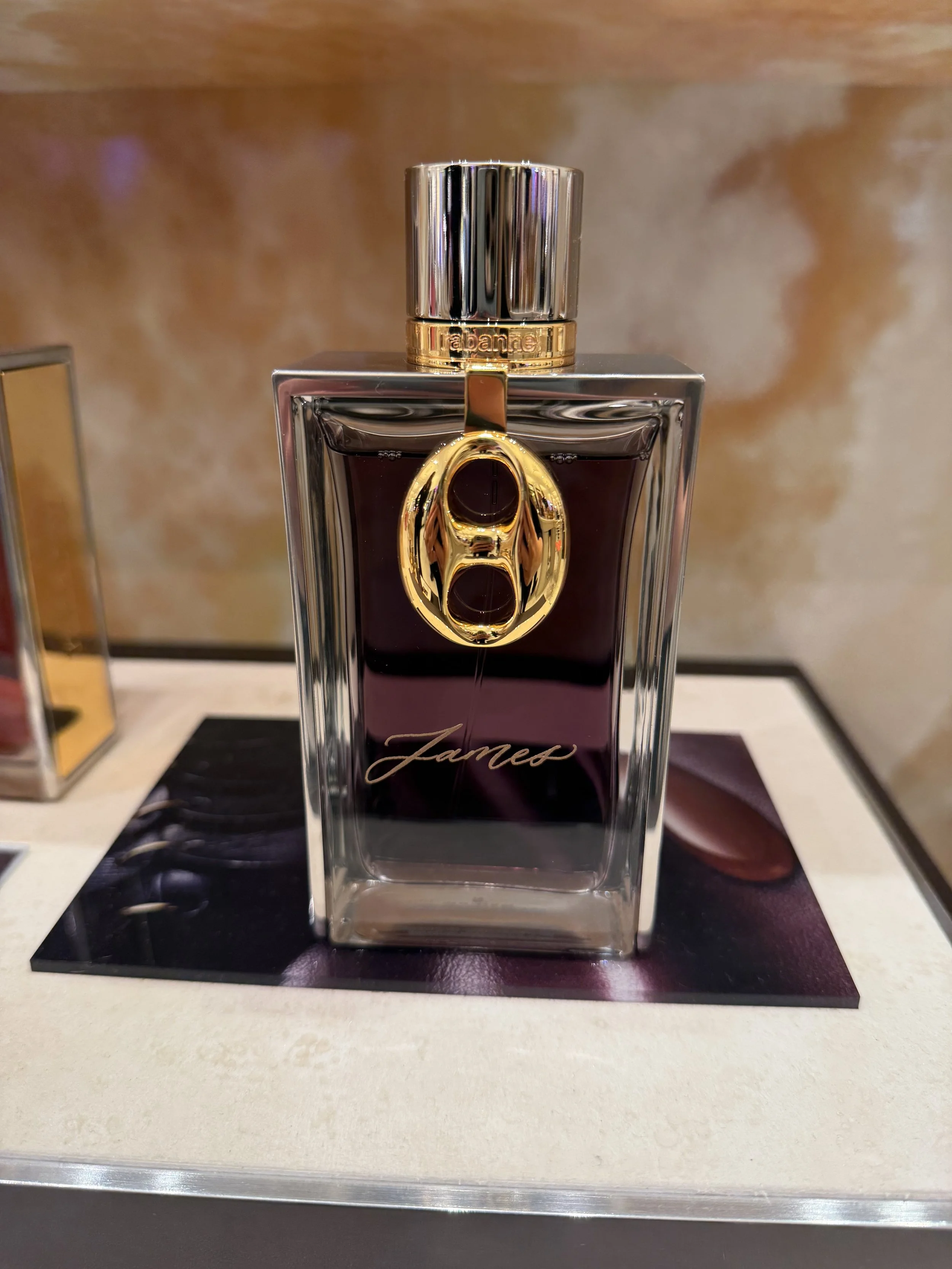 A perfume bottle with a gold and silver cap, a clear rectangular glass body with dark purple liquid inside, gold chain detail, and the name "James" written on the front, displayed on a shelf with a beige and brown background.