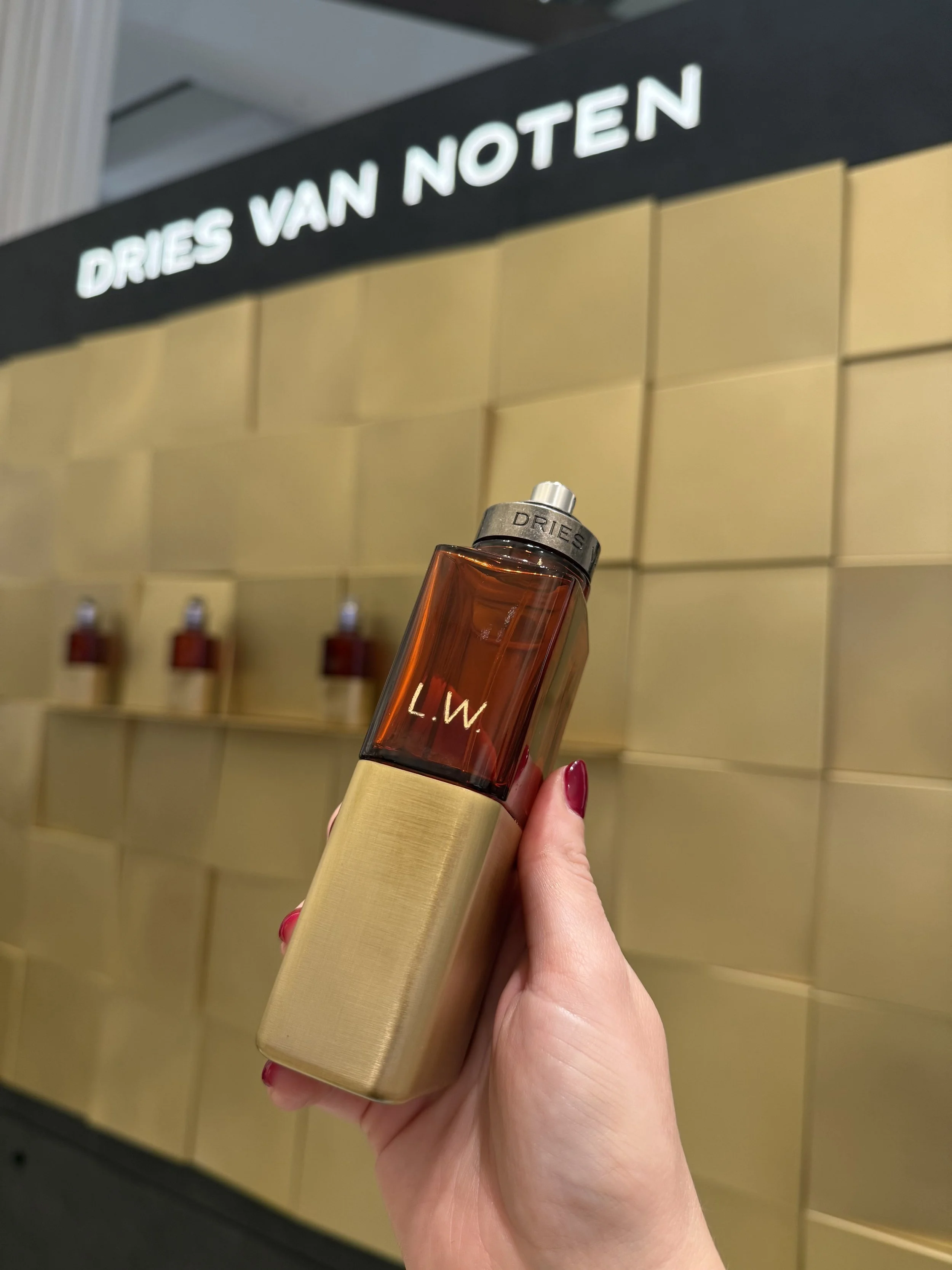 Hand holding a perfume bottle labeled 'L.W' in front of a gold display shelf with other similar bottles, and a sign that says 'DRIES VAN NOTEN' in the background.