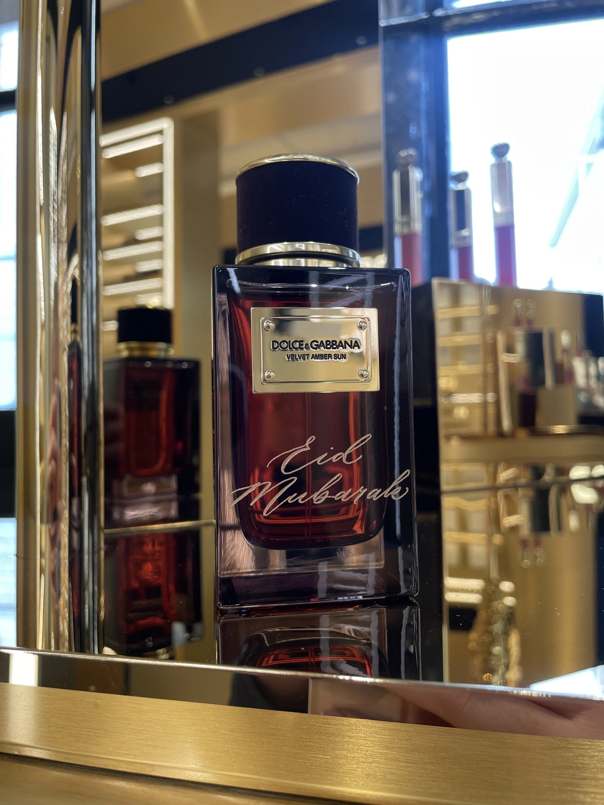 A bottle of Dolce & Gabbana Velvet Amber Sun perfume titled Eid Mubarak on a reflective surface with other perfume bottles and a window in the background.