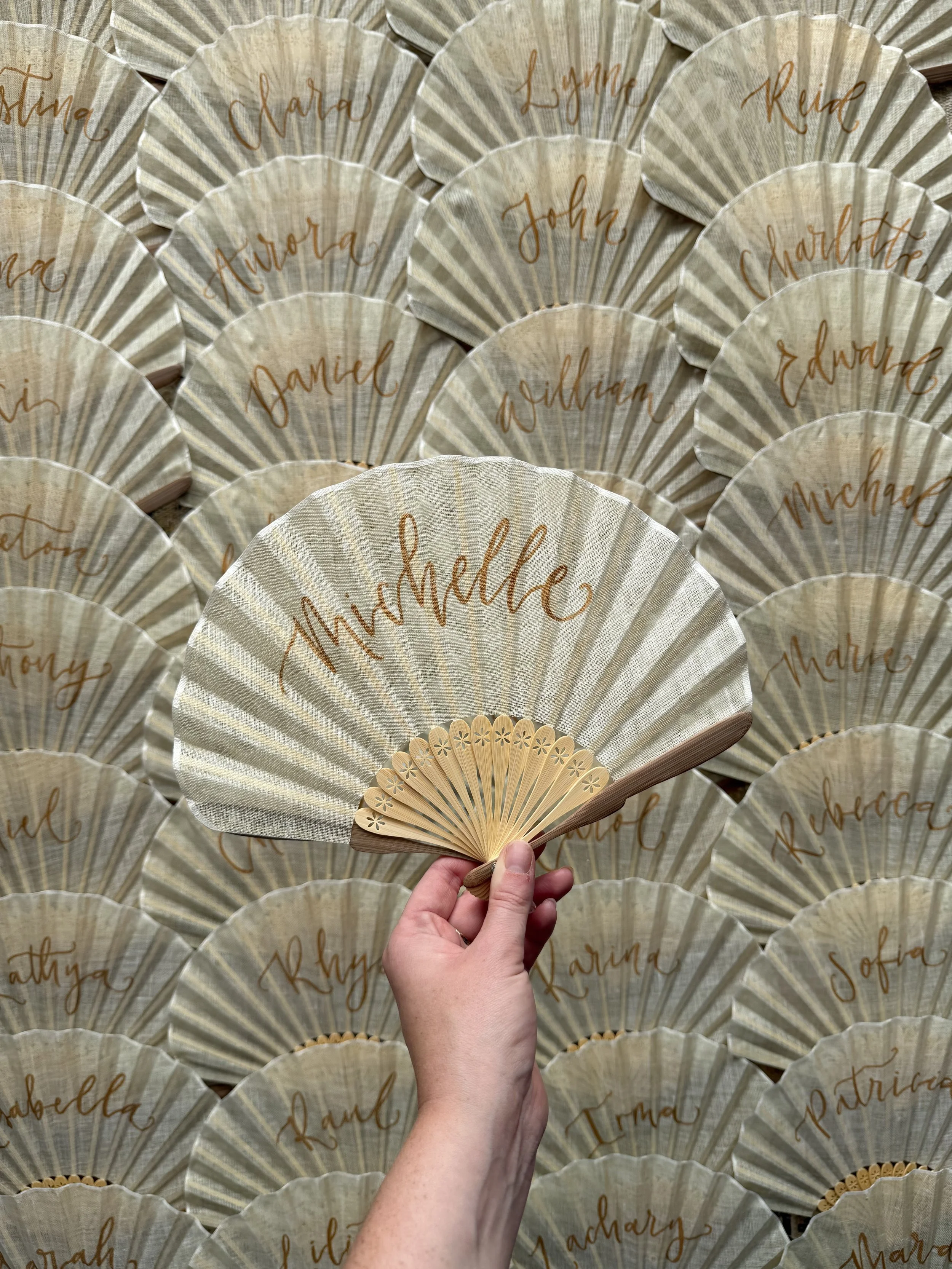 The image shows a hand holding a beige folding fan with the name 'Michelle' written on it in gold lettering. The background consists of many similar fans, each with different names handwritten in gold, such as 'Aurora', 'Daniel', and 'John', arranged