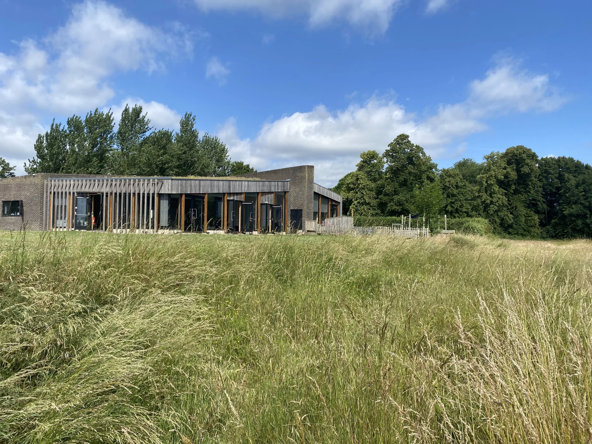 A modern building with wooden accents surrounded by tall grass and trees under a partly cloudy blue sky.