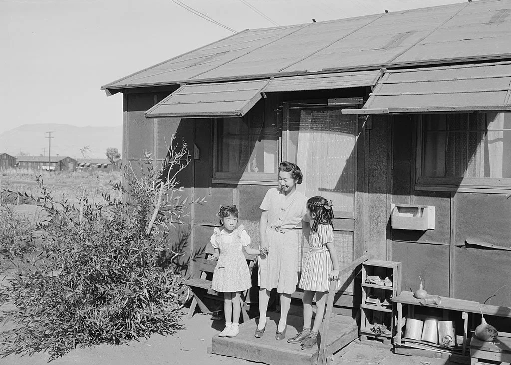 Mrs. Yaeko Nakamura and her two children, Joyce Yuki (right) and Louise Tami (left), standing on the step at the entrance of a dwelling, Manzanar Relocation Center