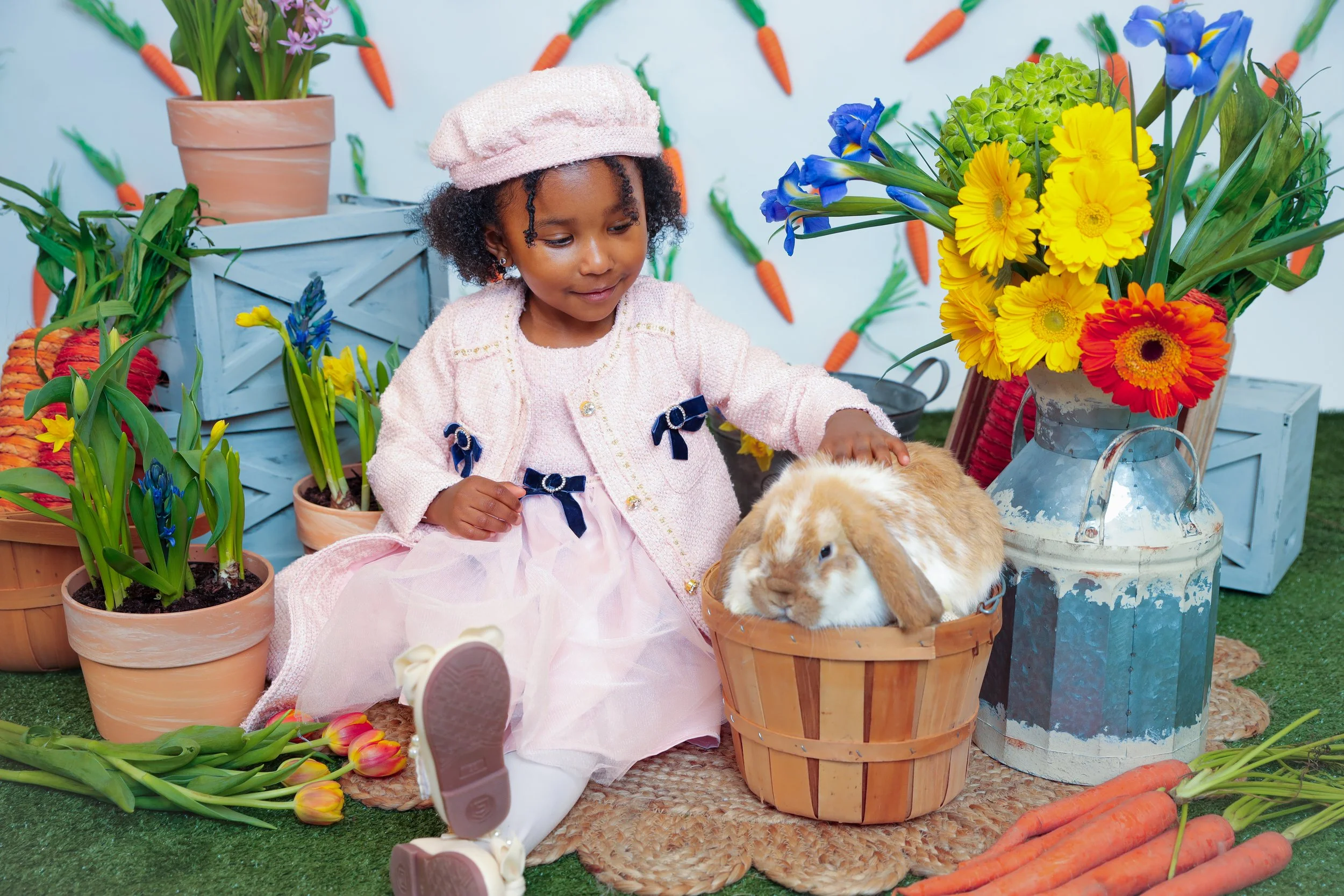 A young girl in pink dress and cardigan, wearing a pink hat, sitting with a brown and white rabbit in a basket. The scene includes colorful flowers and carrots, with a backdrop decorated with carrots, creating a spring or Easter-themed setting.
