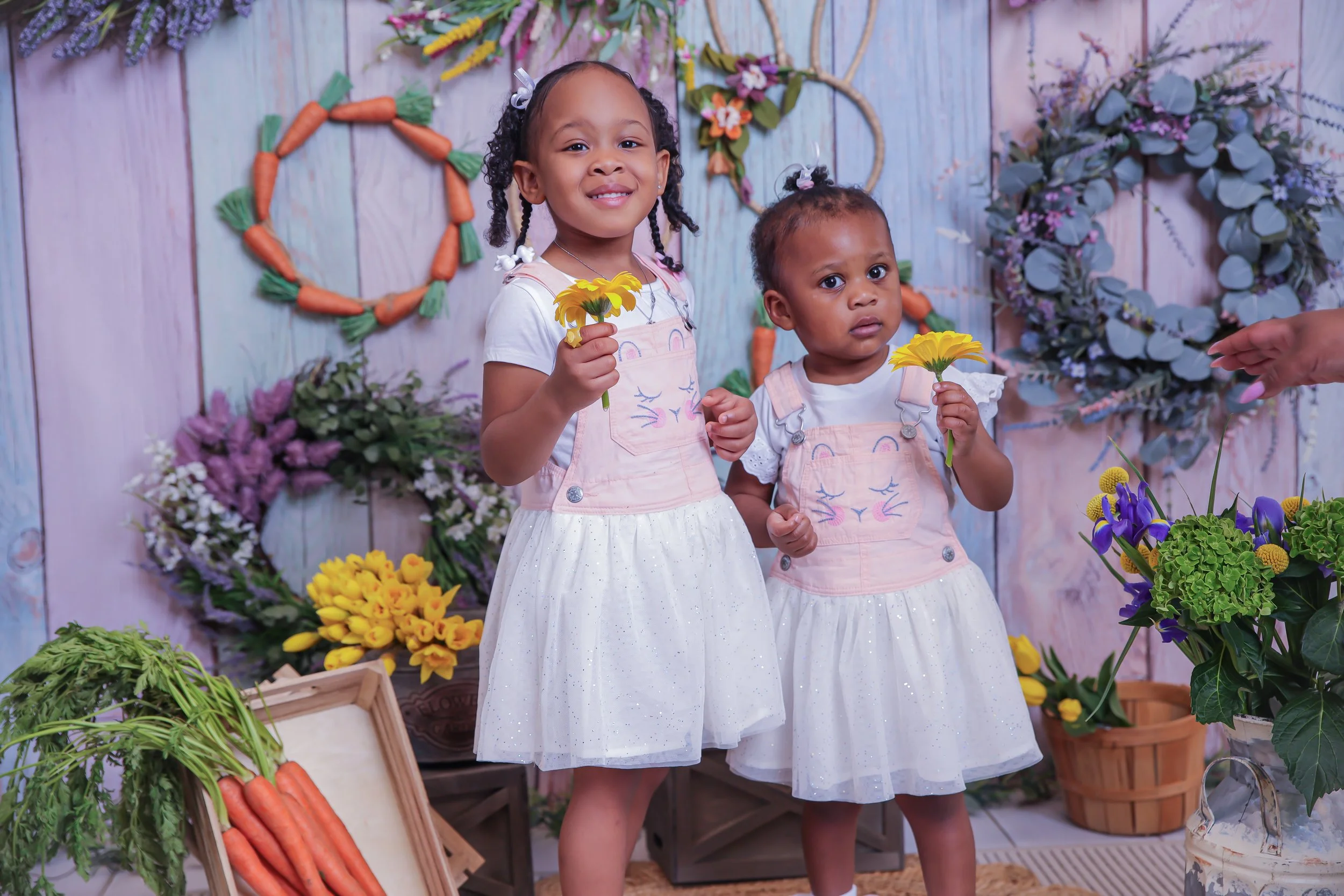 Two young girls holding yellow flowers, standing in front of a decorated pastel-colored floral backdrop with carrot wreaths, flowers, and greenery.