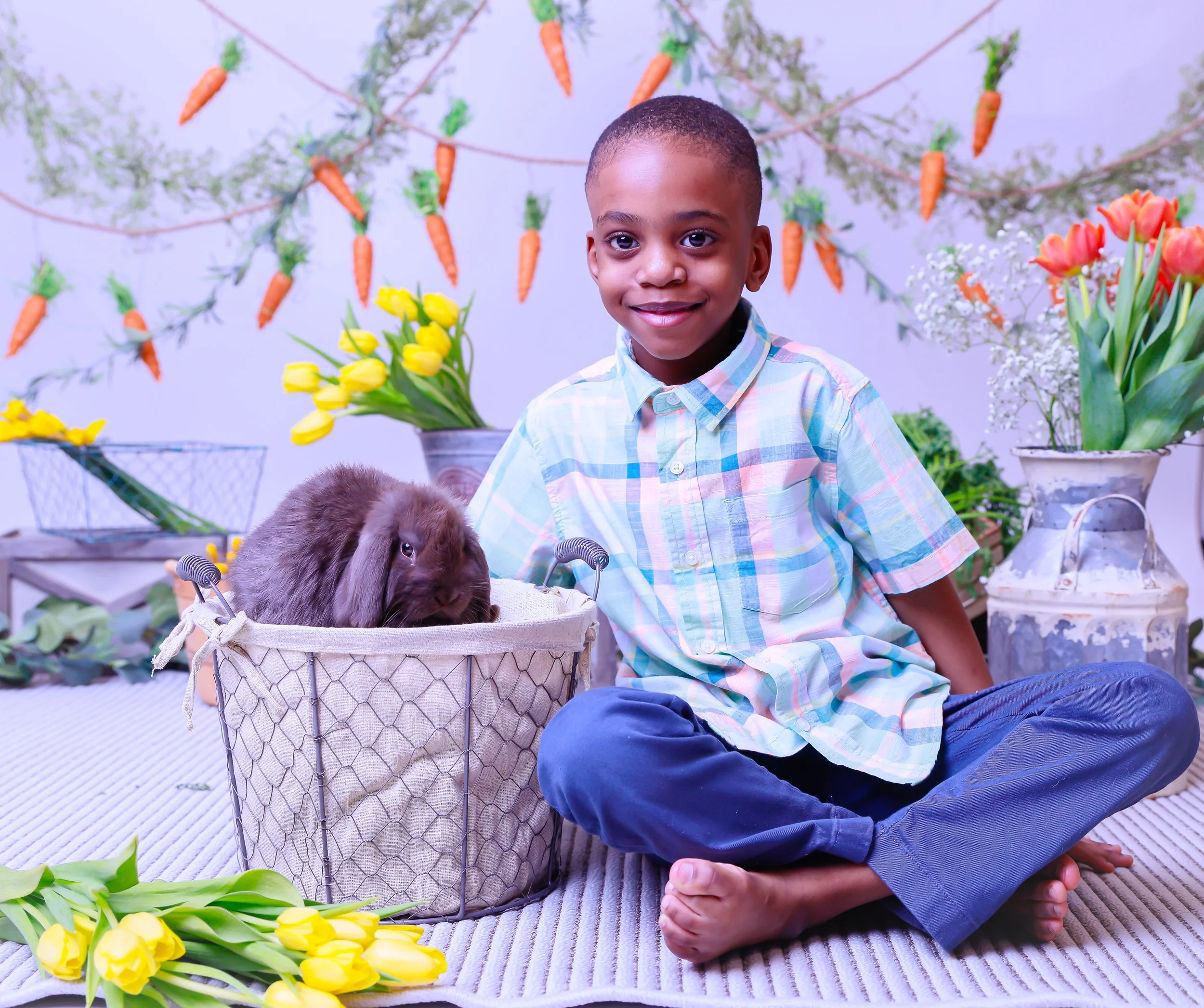 A young boy sitting cross-legged on a striped rug, smiling at the camera, next to a basket with a brown lop-eared rabbit, surrounded by colorful flowers and carrot decorations hanging in the background.
