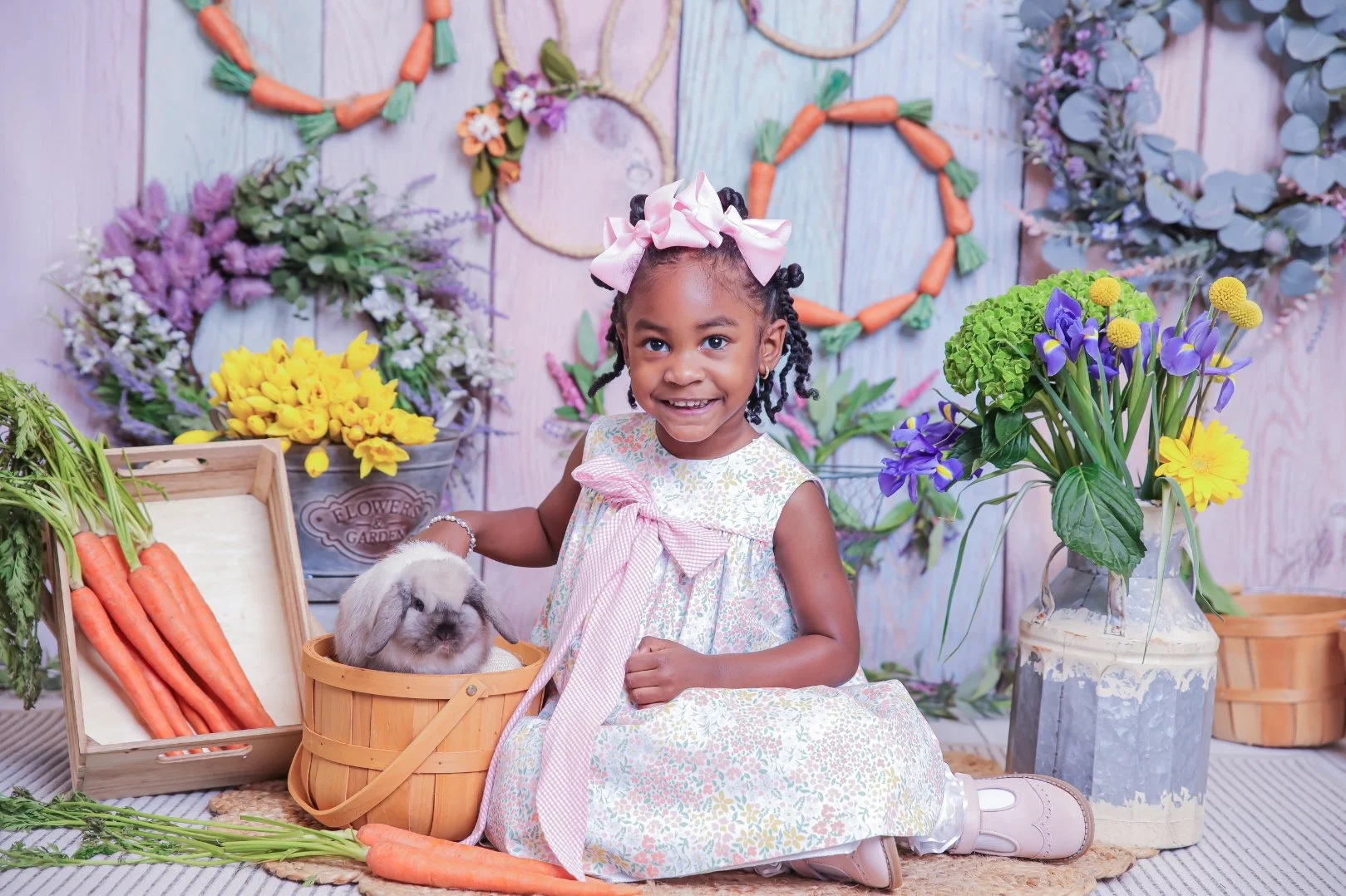 A young girl with braids and a pink bow in her hair, sitting on the floor, holding a basket with a bunny toy, surrounded by carrots and colorful flowers, with a decorated, floral backdrop.