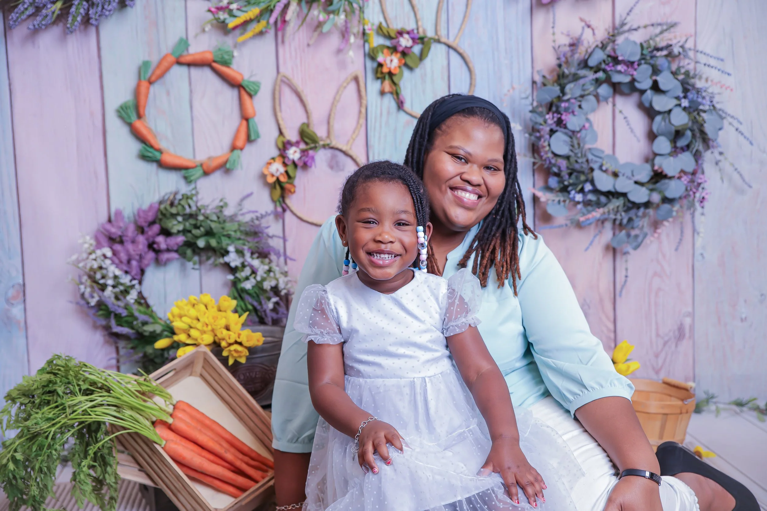A smiling woman and a young girl sitting on a bench surrounded by spring-themed floral decorations and vegetable baskets.