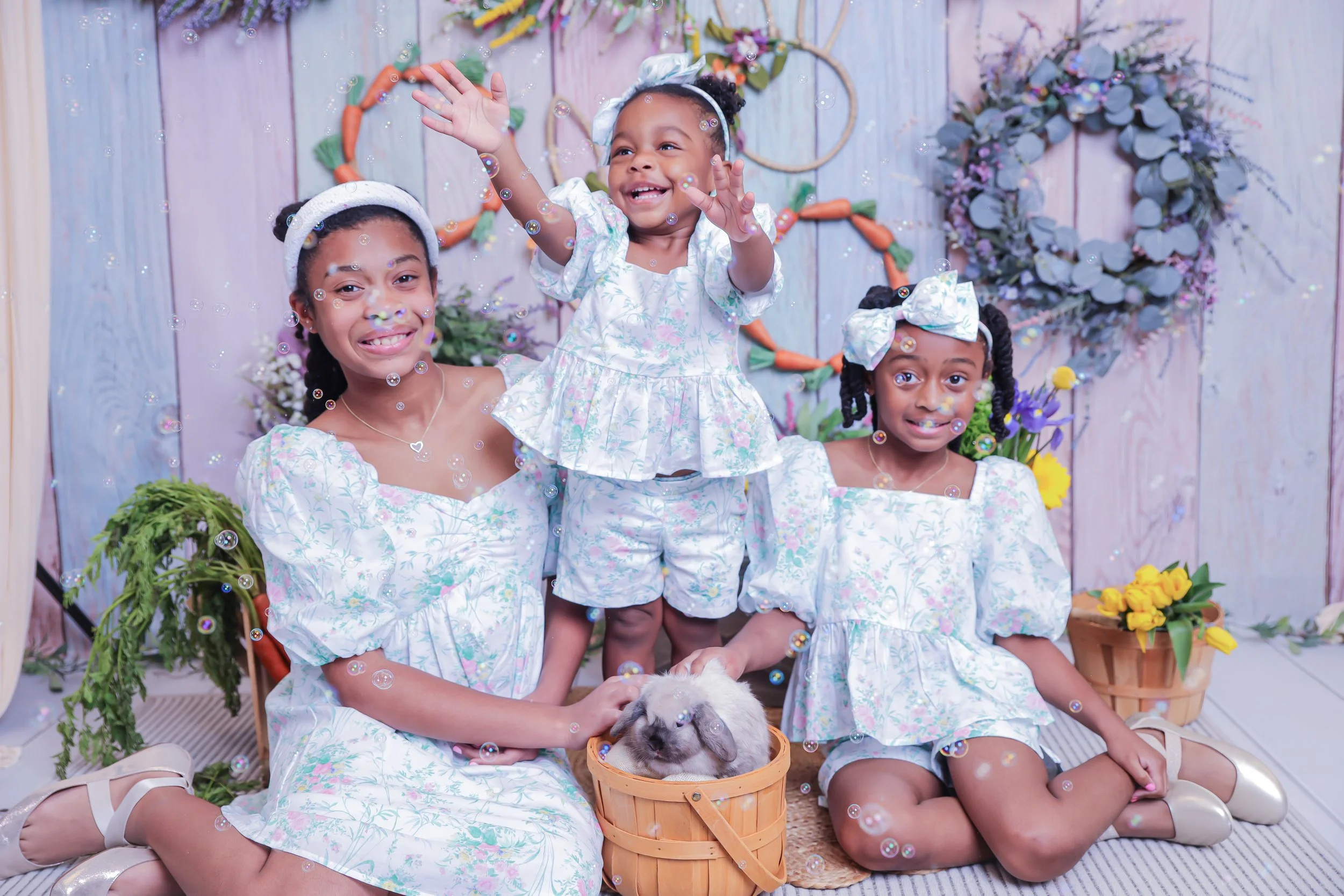 Four young girls with dark curly hair and brown skin, dressed in pastel-colored floral dresses, posing with a small bunny in a wicker basket, surrounded by flowers, greenery, and decorative wreaths, in a spring-themed photo setup with bubbles floatin