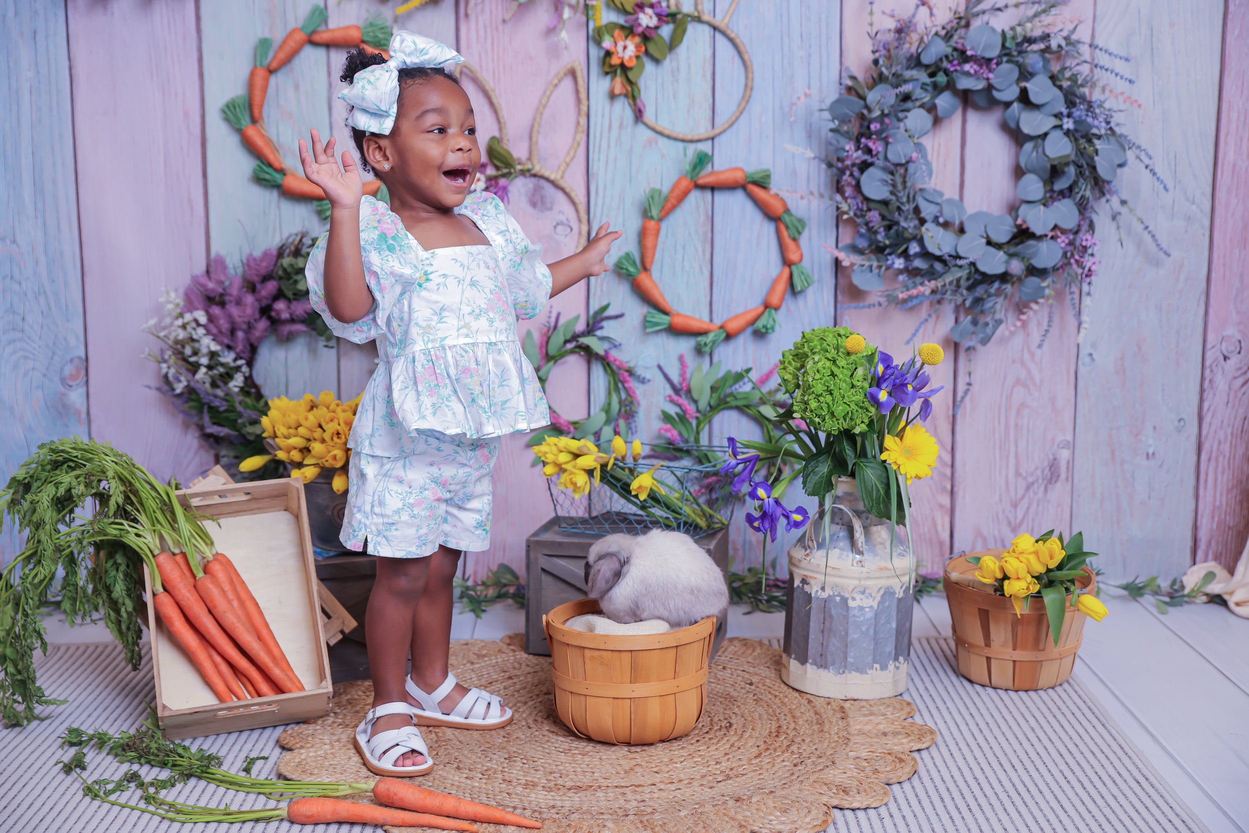 A young girl standing in a flower shop, smiling and waving, surrounded by colorful flowers, flower wreaths, and a rabbit sitting in a basket.