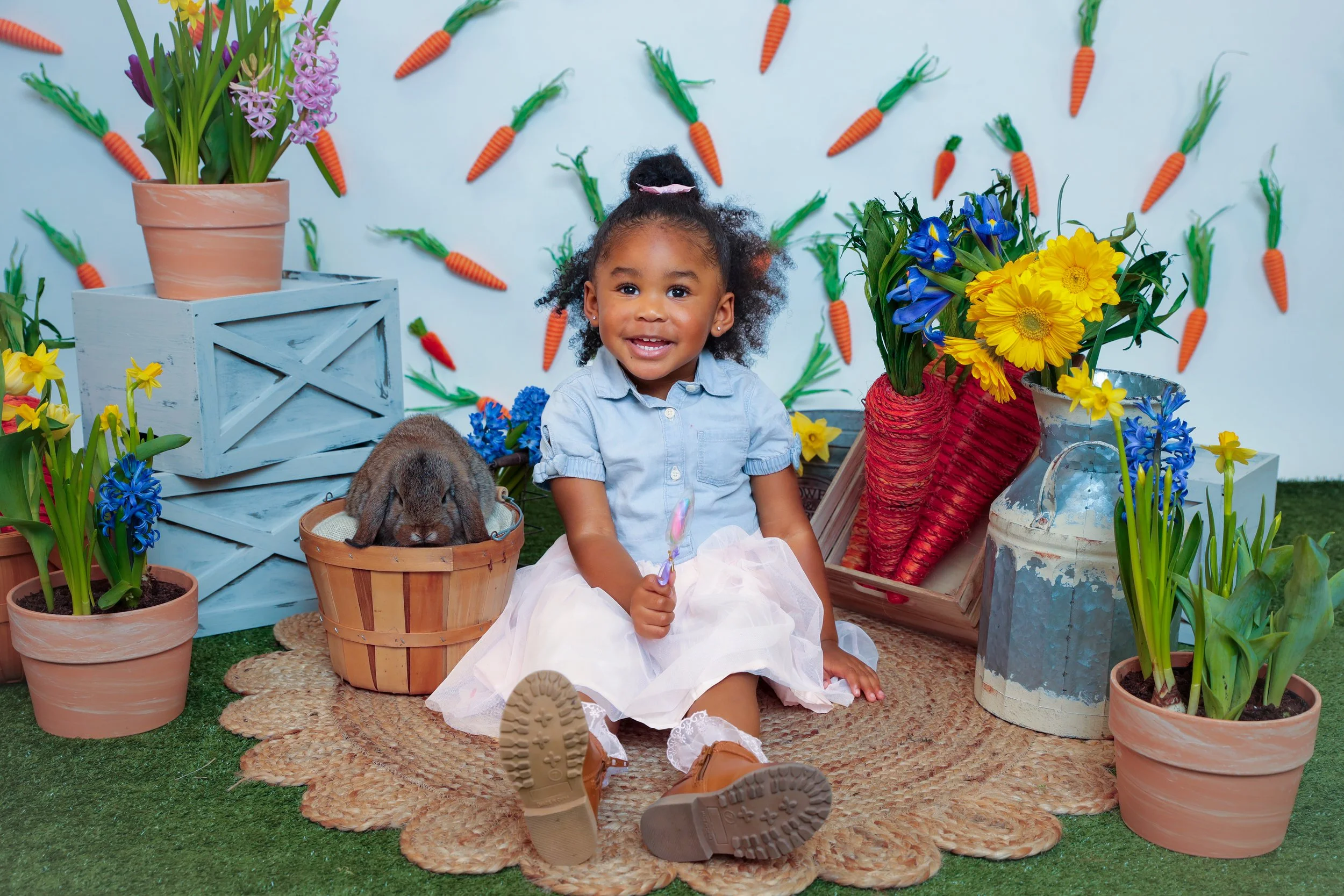 A smiling young girl sitting on a woven rug surrounded by colorful flowers and potted plants. A small bunny is in a basket next to her, with paper carrots on the wall in the background.