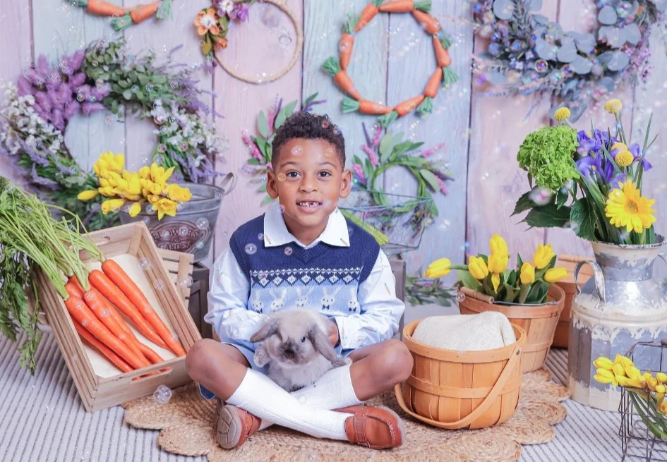 Young boy sitting on the floor holding a rabbit, surrounded by colorful spring flowers and vegetable decorations, with flower wreaths and garden-themed decor in the background.