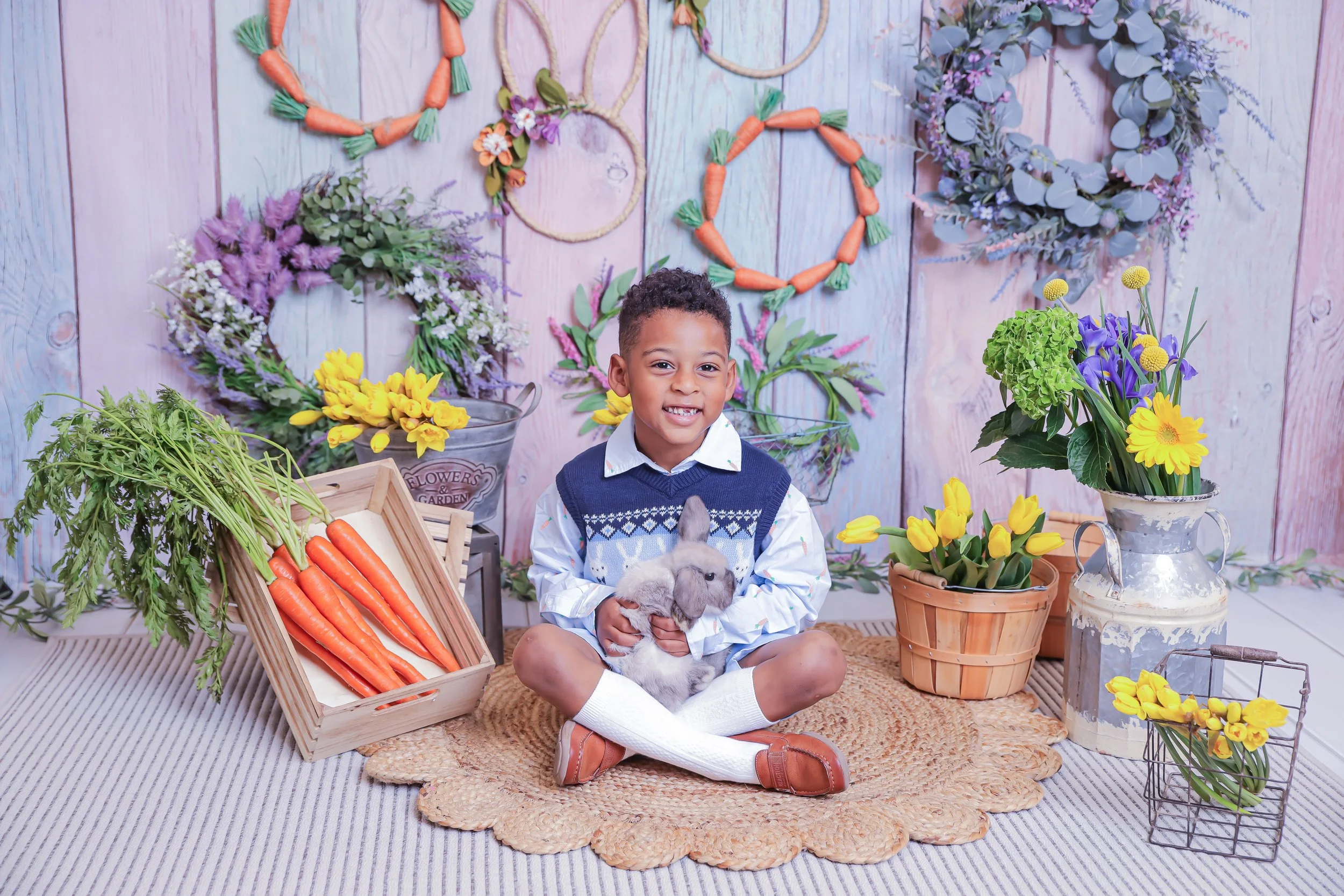 A young boy sitting on a round rug holding a gray rabbit with a background of colorful floral wreaths and pots of flowers, including carrots, sitting on woven and metal containers.