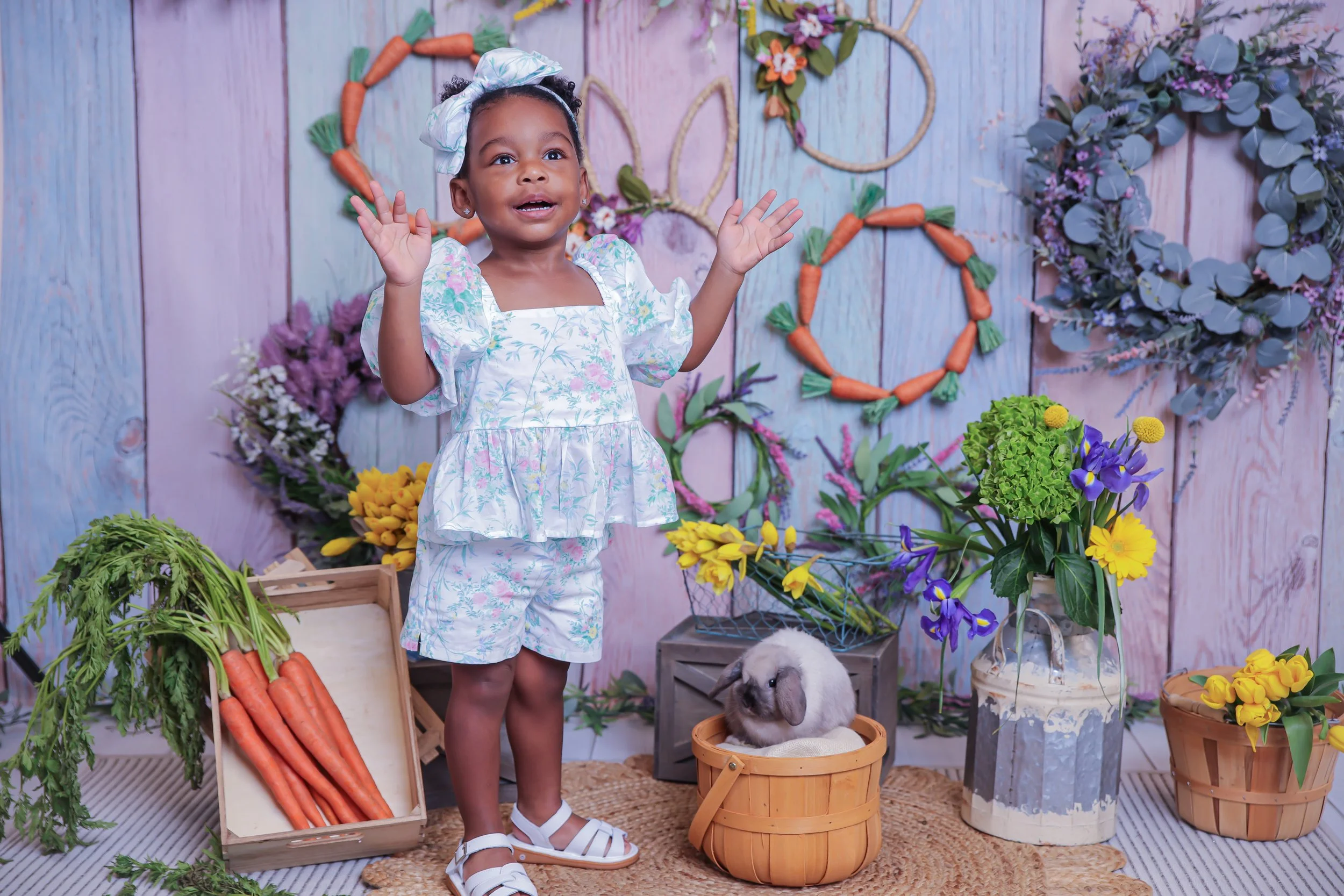 A young girl in pastel floral pajamas and sandals stands with her hands raised, smiling in a colorful flower and vegetable farm scene. There are carrots in a basket, various flowers in pots and in the background, and a small rabbit sitting in a baske