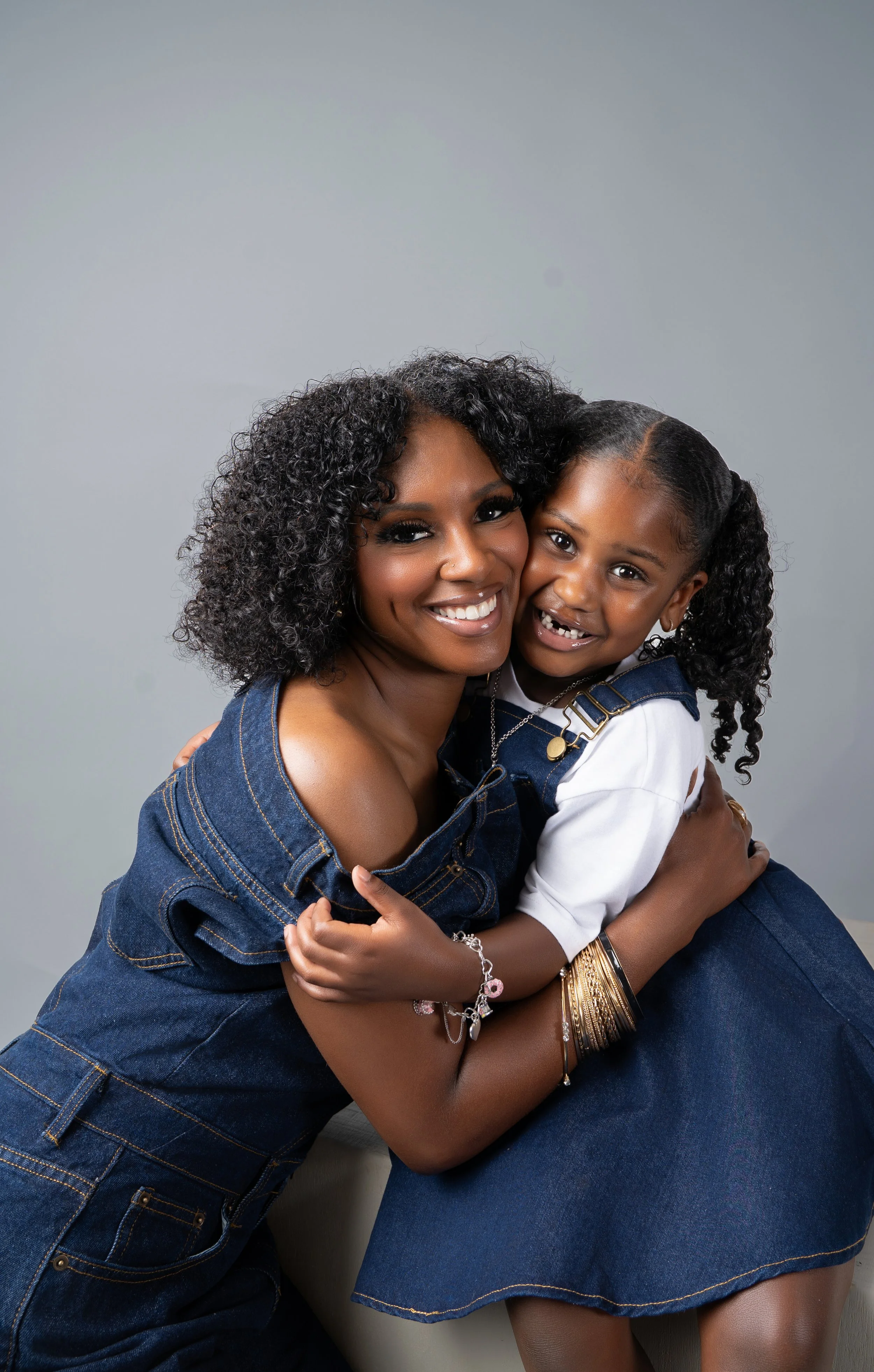 An African American woman and a young girl hugging each other and smiling, both wearing denim outfits, in front of a plain gray background.
