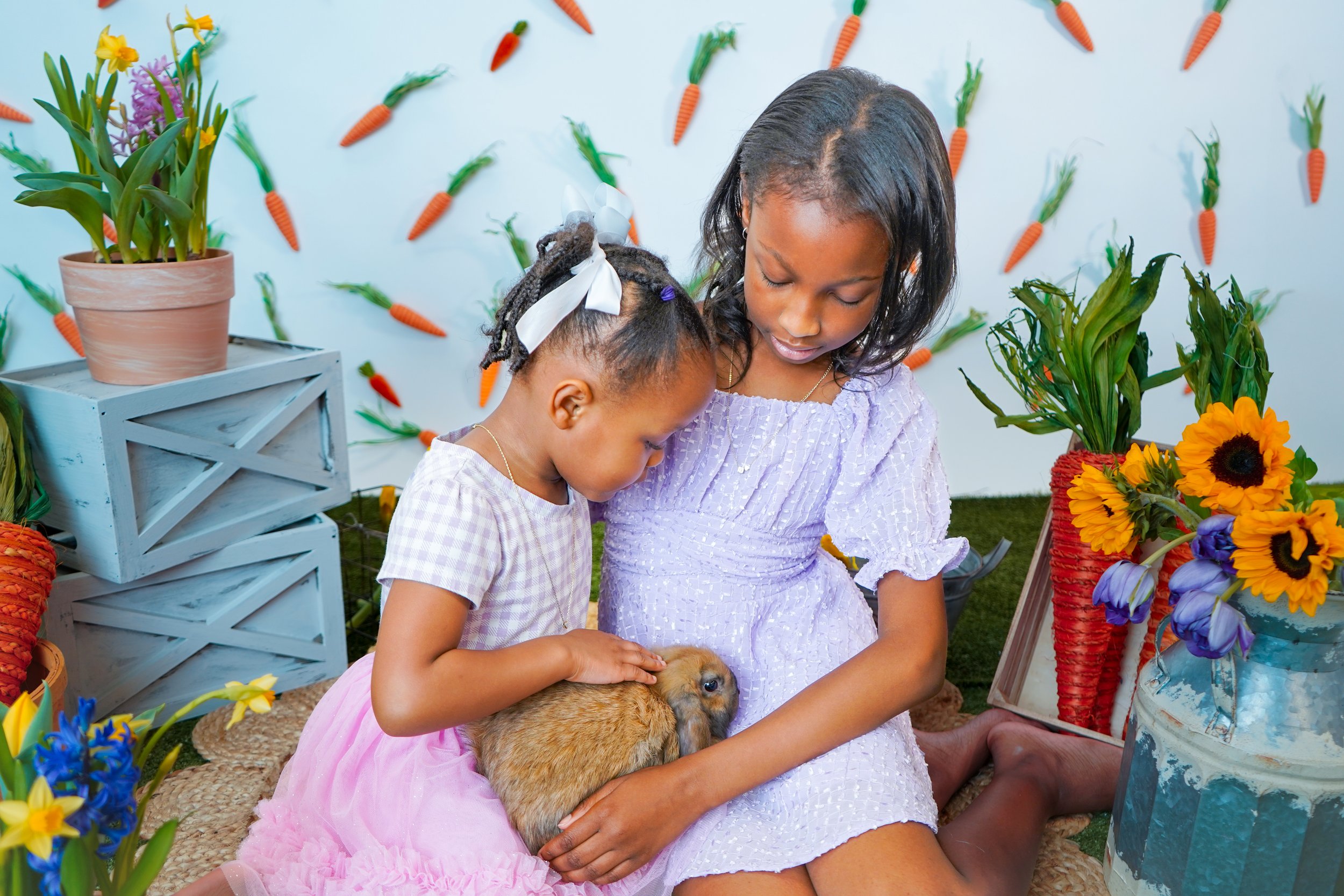 Two young girls sitting on a woven mat, holding and petting a brown rabbit, amidst colorful floral decorations and potted plants, with carrots on the wall behind them.