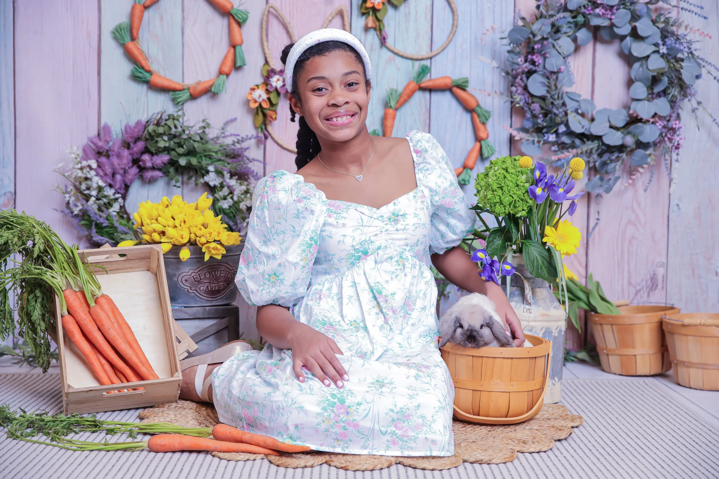 Young girl sitting on the floor with a bunny inside a basket, surrounded by colorful flowers and carrots in a garden-themed setting.