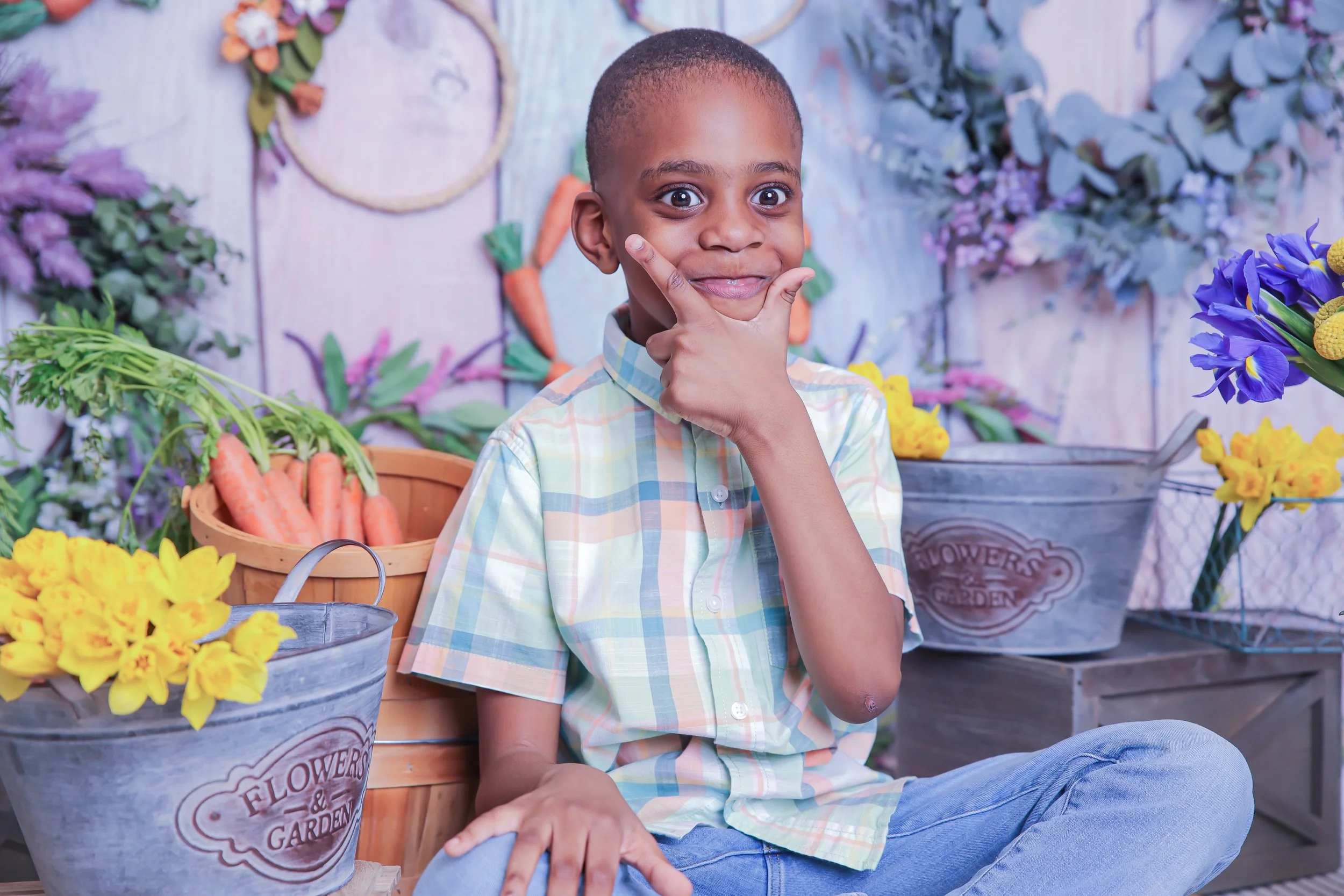 Young boy sitting among flower and vegetable baskets at a flower garden, making a thinking face with his hand on his chin.