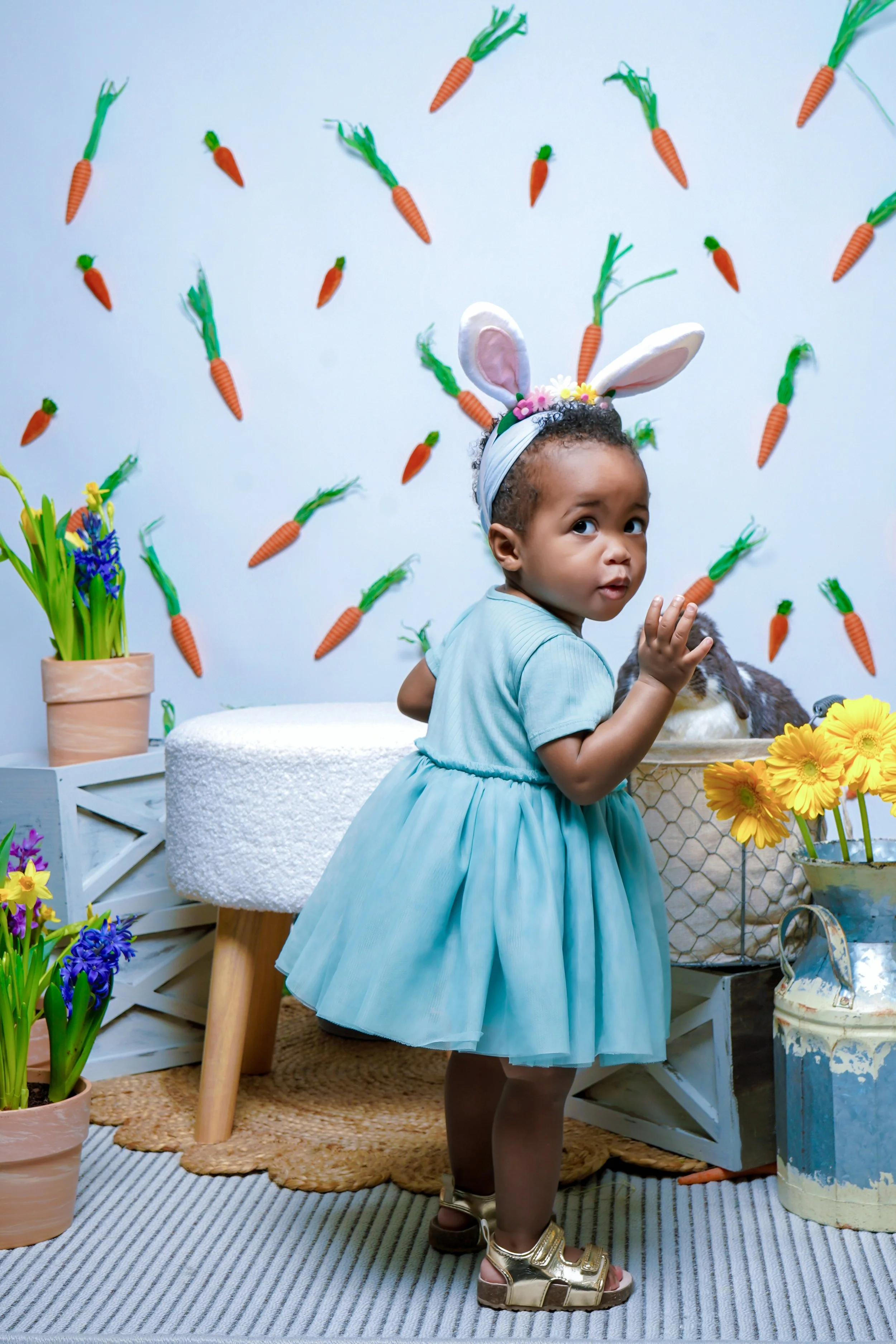 A young girl dressed as a bunny in a light blue dress and bunny ears headband, standing in a decorated setting with carrot and flower decorations.