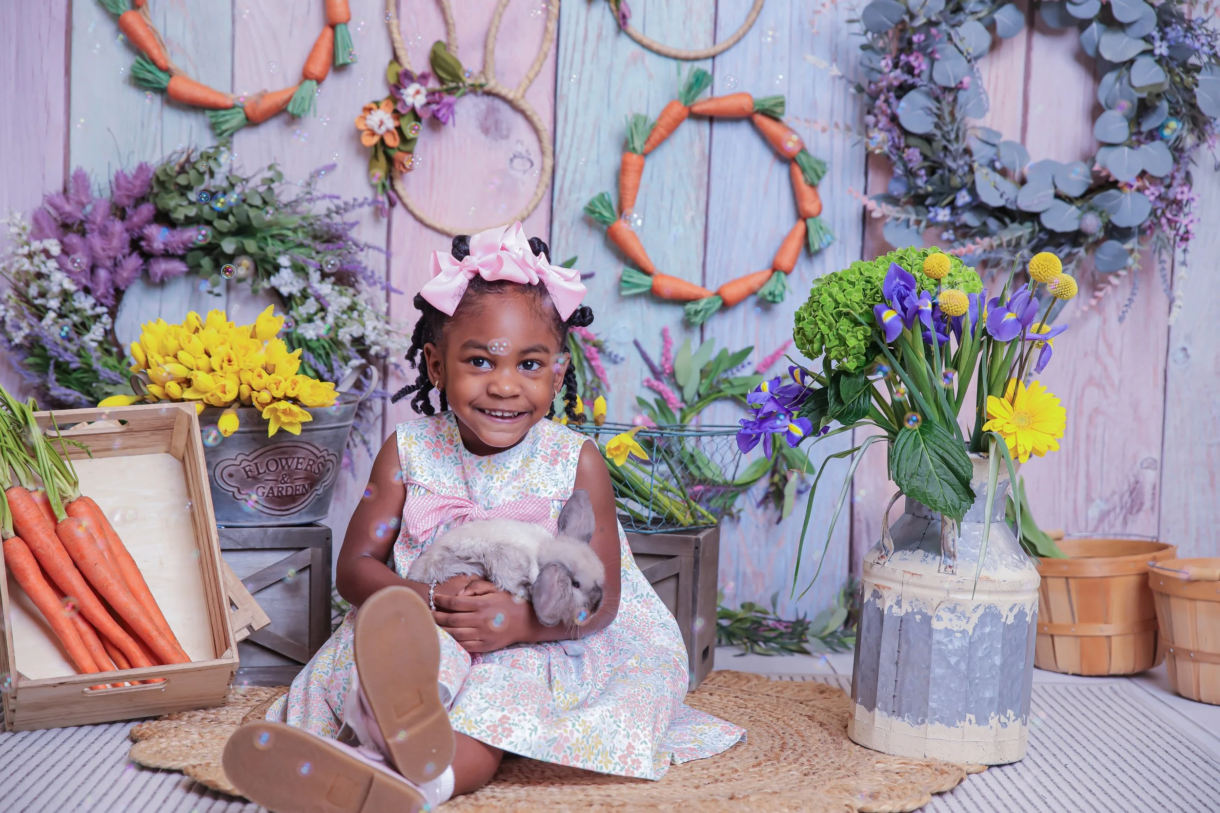 A young girl with a pink bow in her hair is sitting on a woven rug, holding a lop-eared rabbit. Behind her are colorful flower arrangements and wreaths, with a light wooden wall decorated with more wreaths and floral ornaments. The setting resembles 