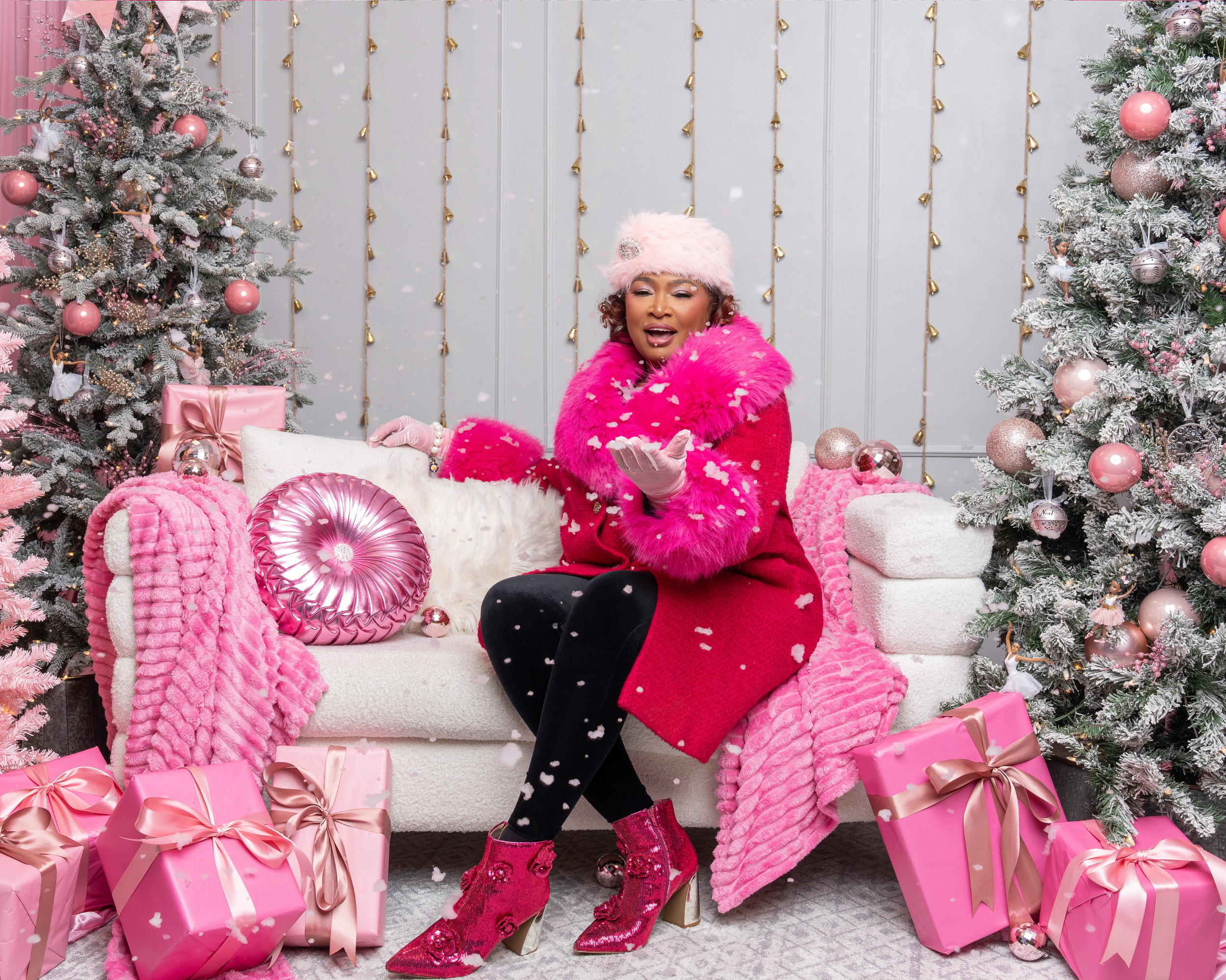 A woman dressed in a pink and black winter holiday outfit sits on a cream-colored sofa, surrounded by pink and white Christmas decorations, including trees, wrapped gifts, and plush blankets.
