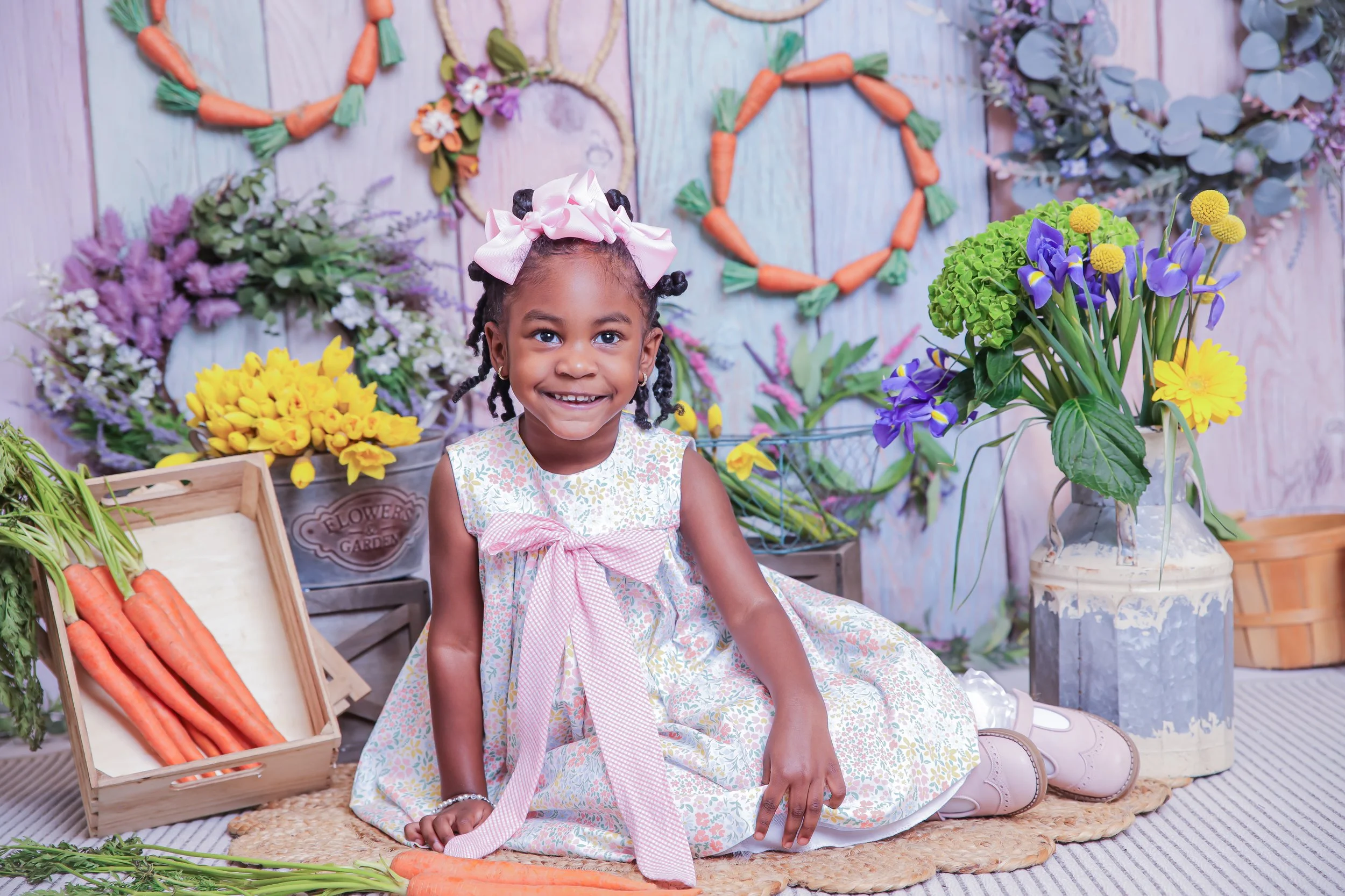A young girl with braided hair and a pink bow headband sitting on a woven mat surrounded by colorful flowers and vegetables in a bright, decorated setting.