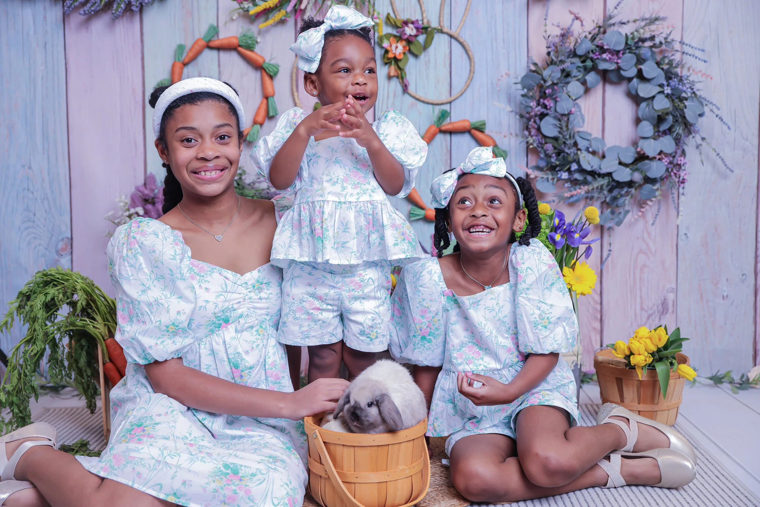 Three young girls in matching floral dresses with puffed sleeves and bows in their hair, smiling and posing indoors with a bunny in a basket, surrounded by colorful flowers and floral wreaths on a pastel wooden backdrop.
