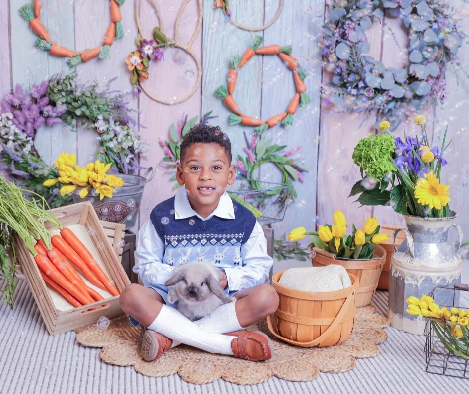 A young boy sitting cross-legged on a woven rug, holding a rabbit, surrounded by spring flowers and carrots, with floral wreaths on the wall behind him.