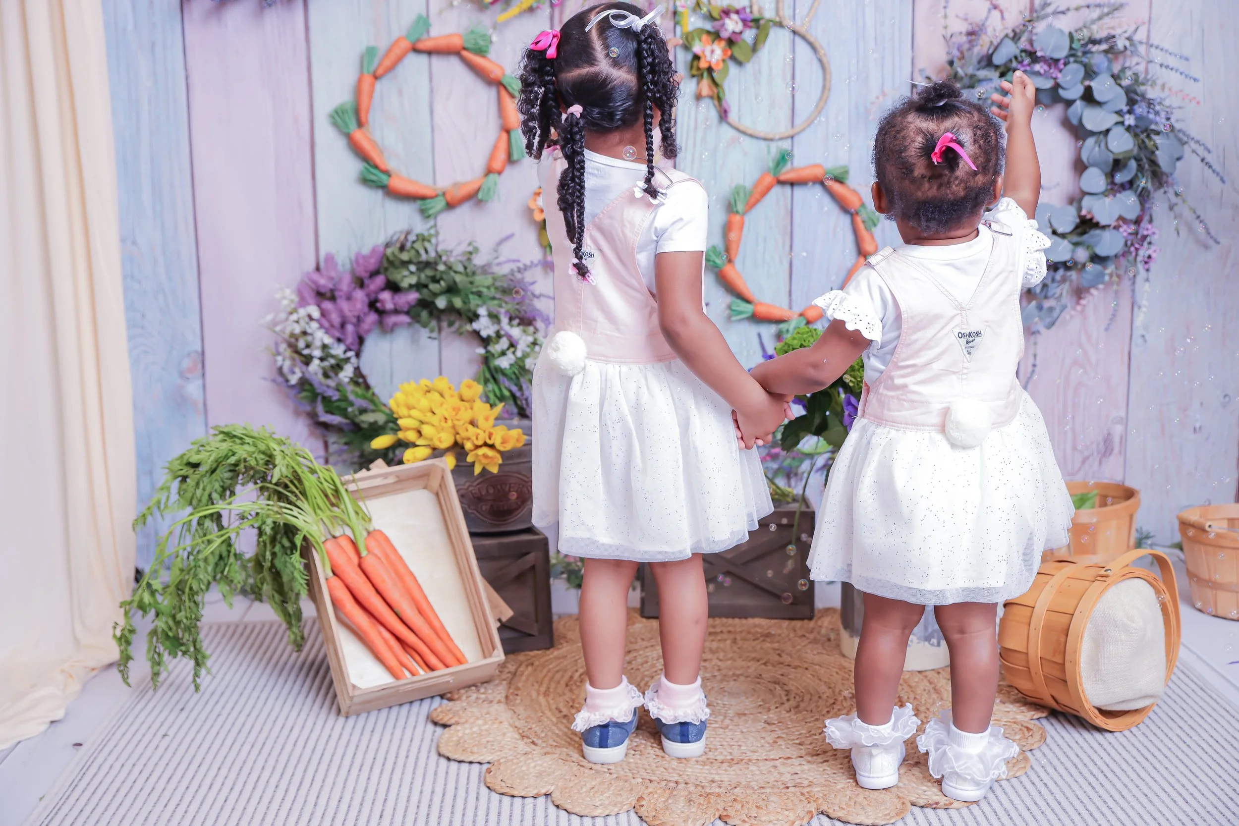 Two young girls holding hands, facing a floral and carrot-themed wall display with wreaths and plant baskets, on a woven rug in a decorated room.