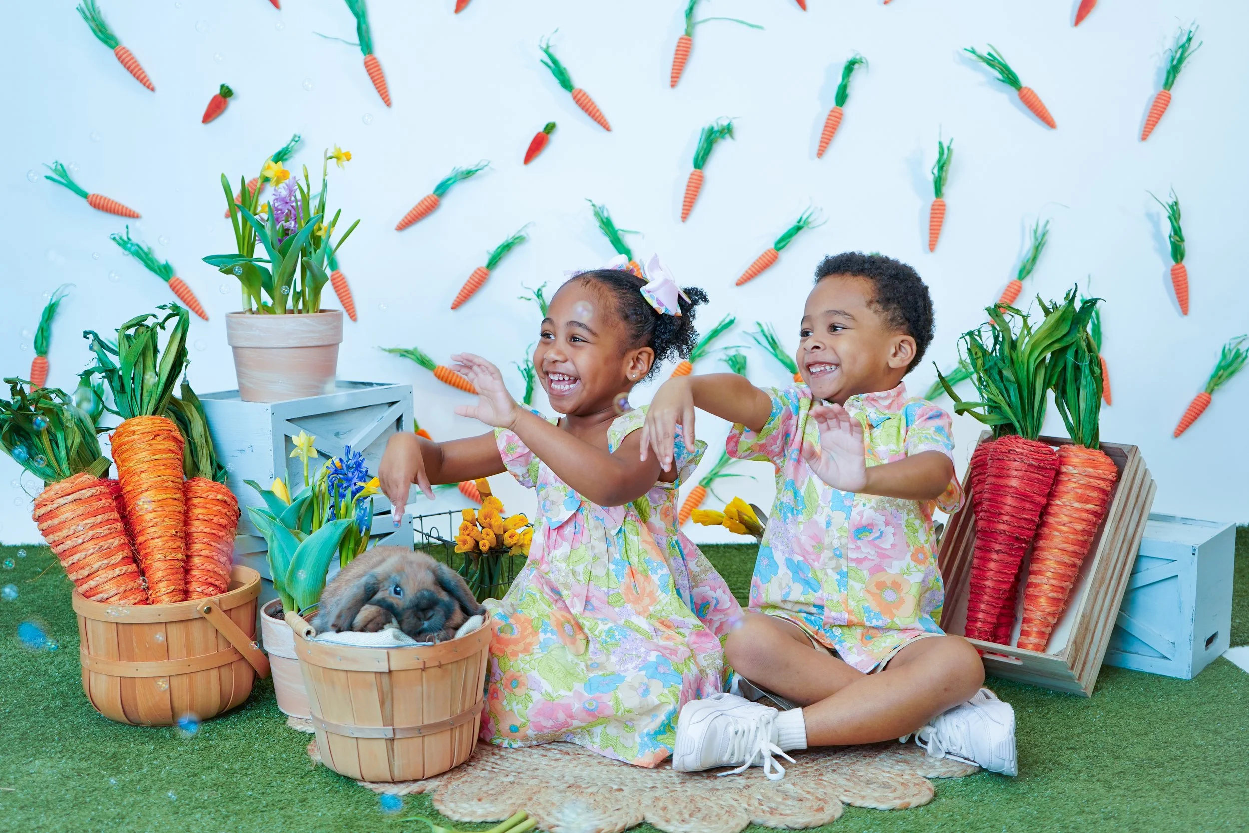 Two children, a girl and a boy, sitting on a rug with Easter-themed decorations. The girl has a bow in her hair and is smiling, while the boy is also smiling. Surrounding them are baskets filled with large, decorative carrots, potted tulips, and othe