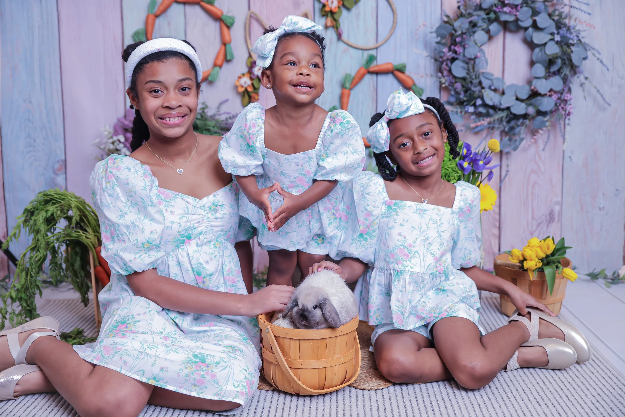 Four young girls in matching floral dresses sitting and kneeling around a rabbit in a basket, with a decorated floral background.