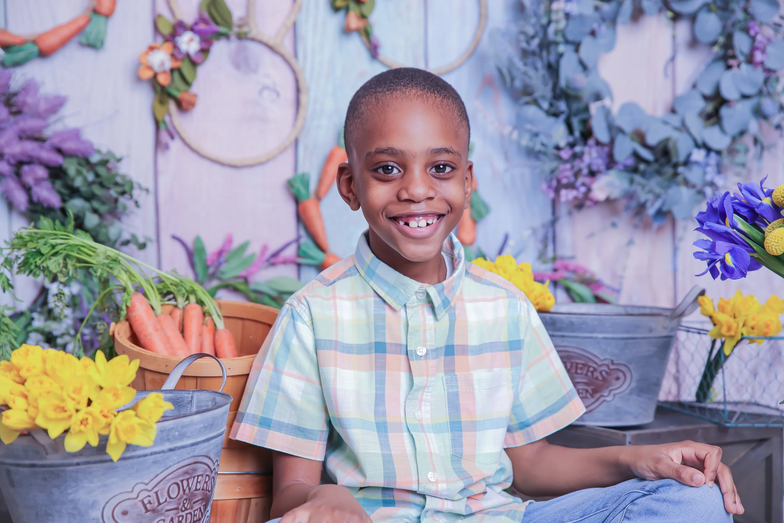 A smiling young boy with missing teeth, sitting in front of flowers and carrots in a rustic setting.